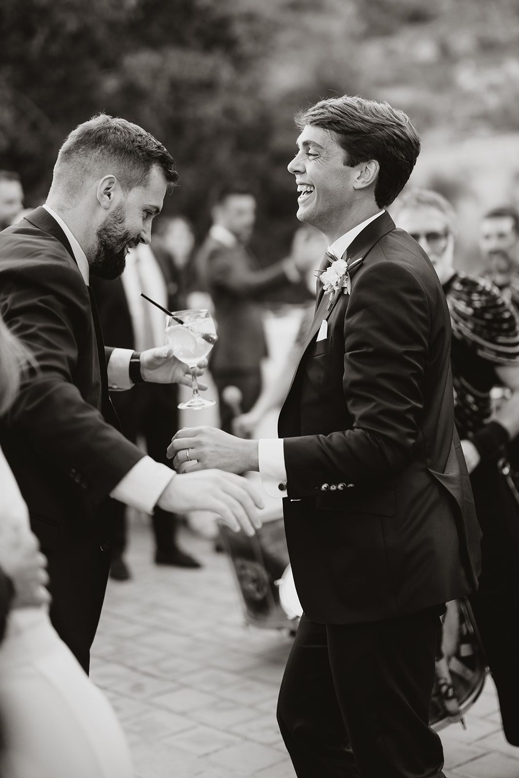 Two wedding guests in suits share a joyful moment of laughter during an outdoor celebration.