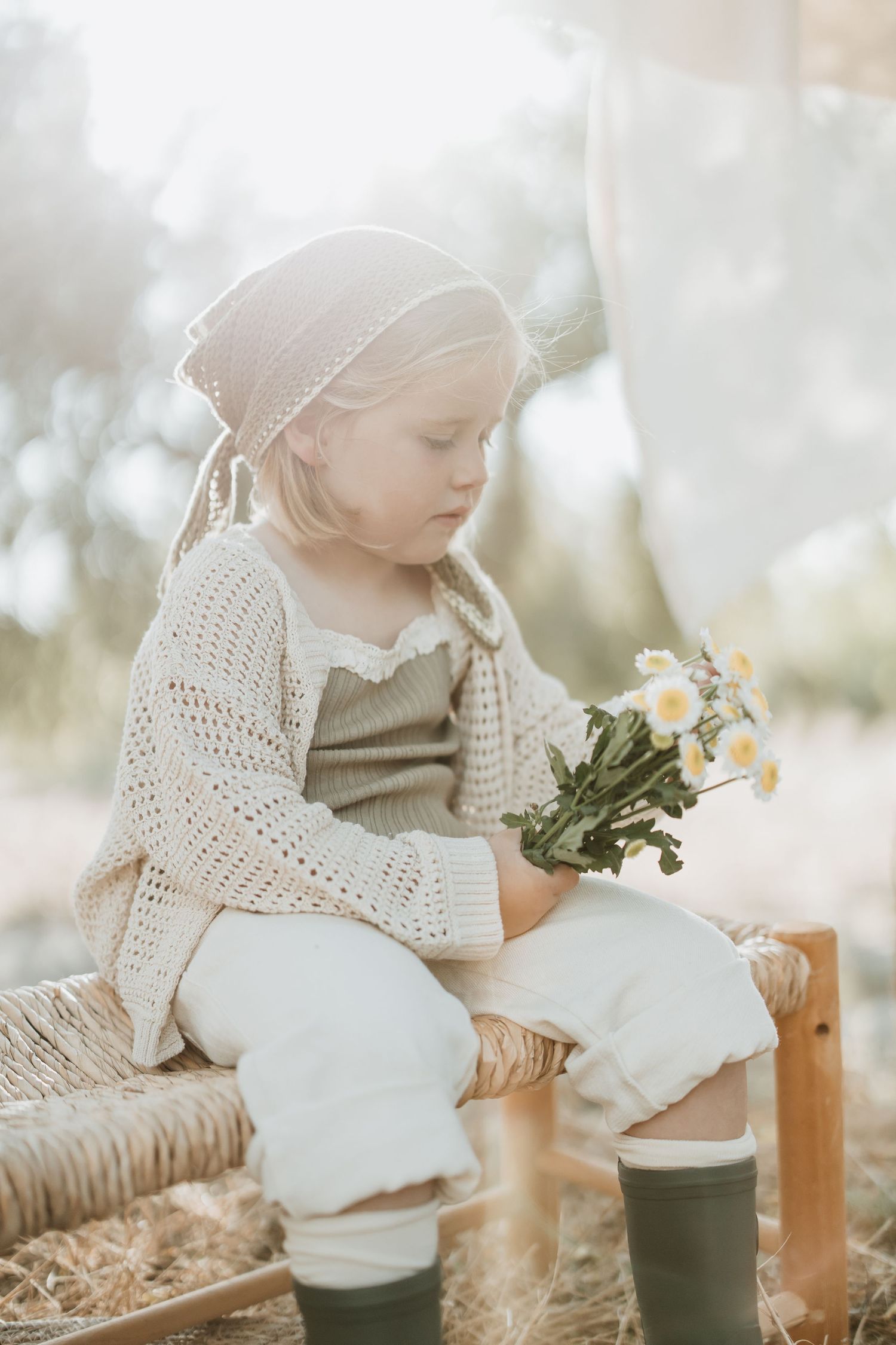 Child in cream knit wear and headscarf examining flowers while seated on woven bench outdoors.