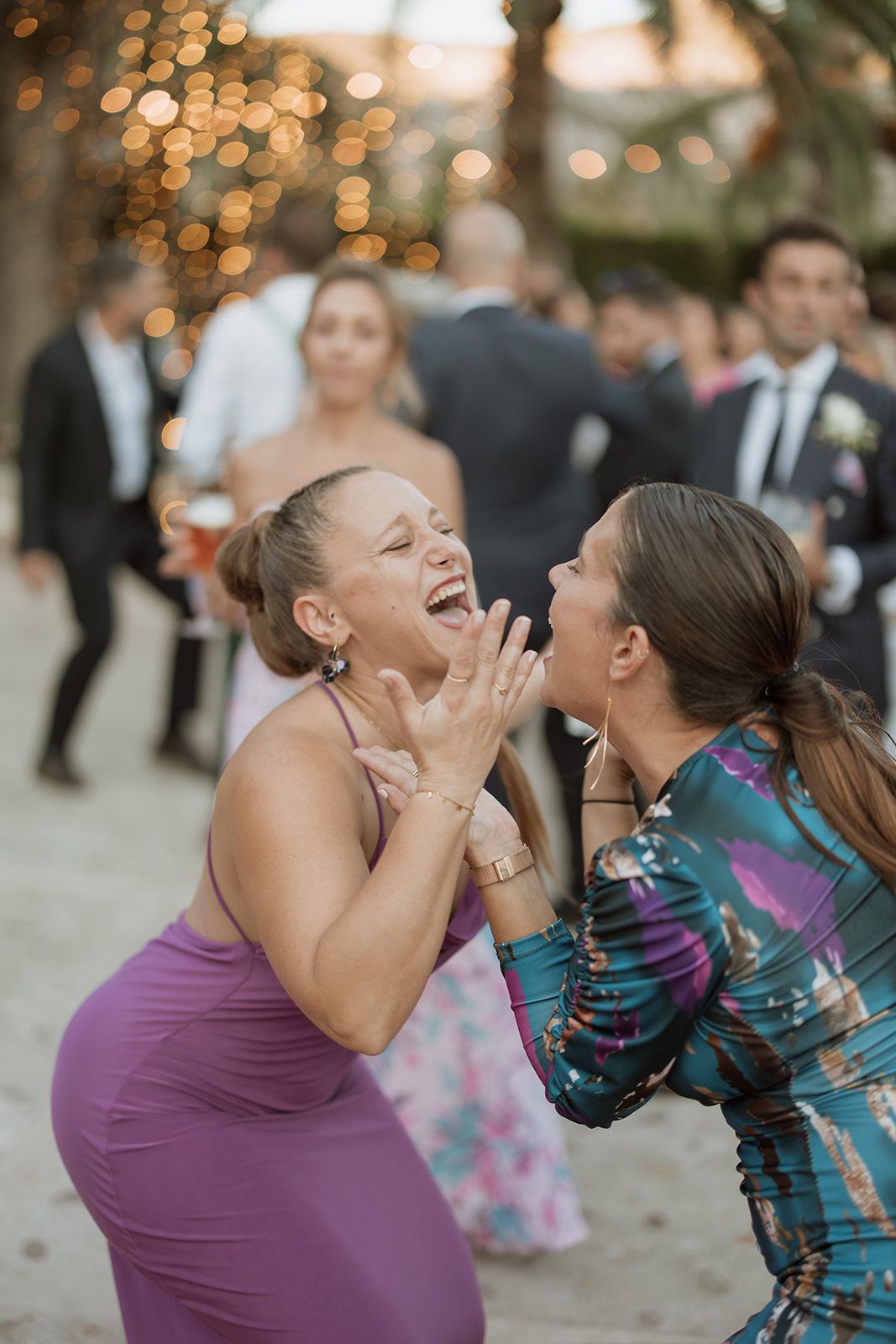 Two wedding guests laughing joyfully together at an outdoor reception with bokeh lights.
