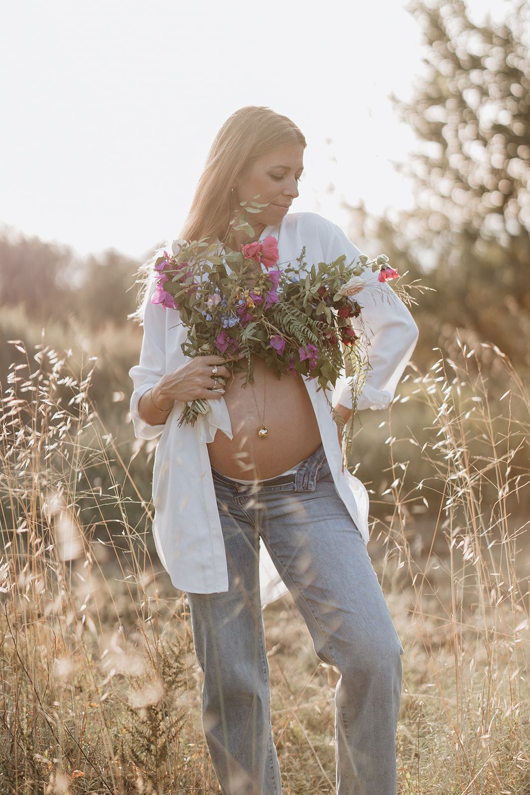 A maternity photo in a wild field with someone holding wildflowers against a sunset backdrop.