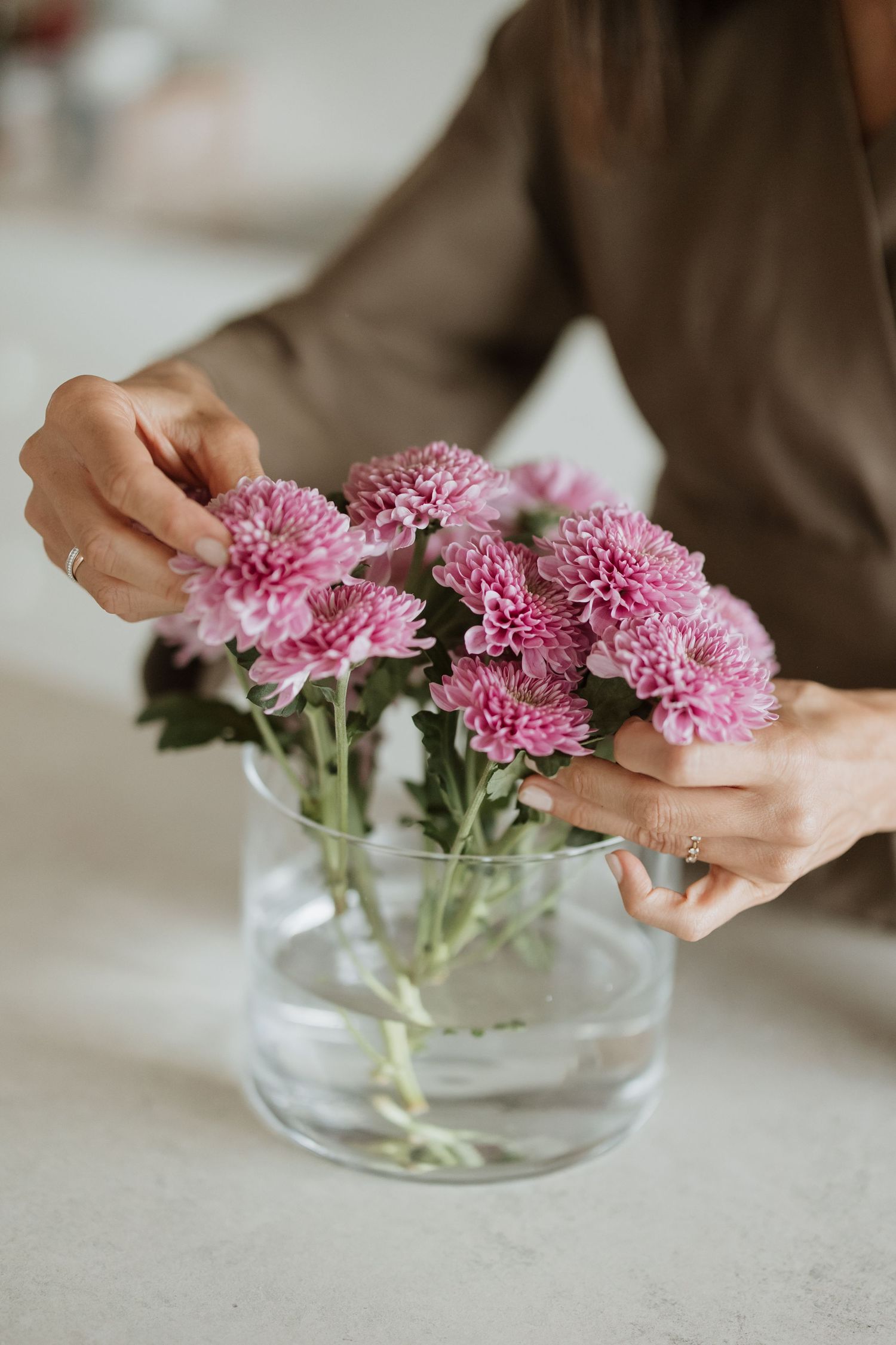 Hands delicately adjusting pink flowers in round glass vase on light surface.