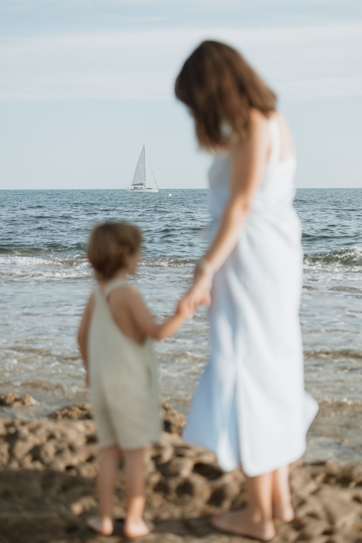 Parent and child watching sailboat on horizon while standing together on beach shore.