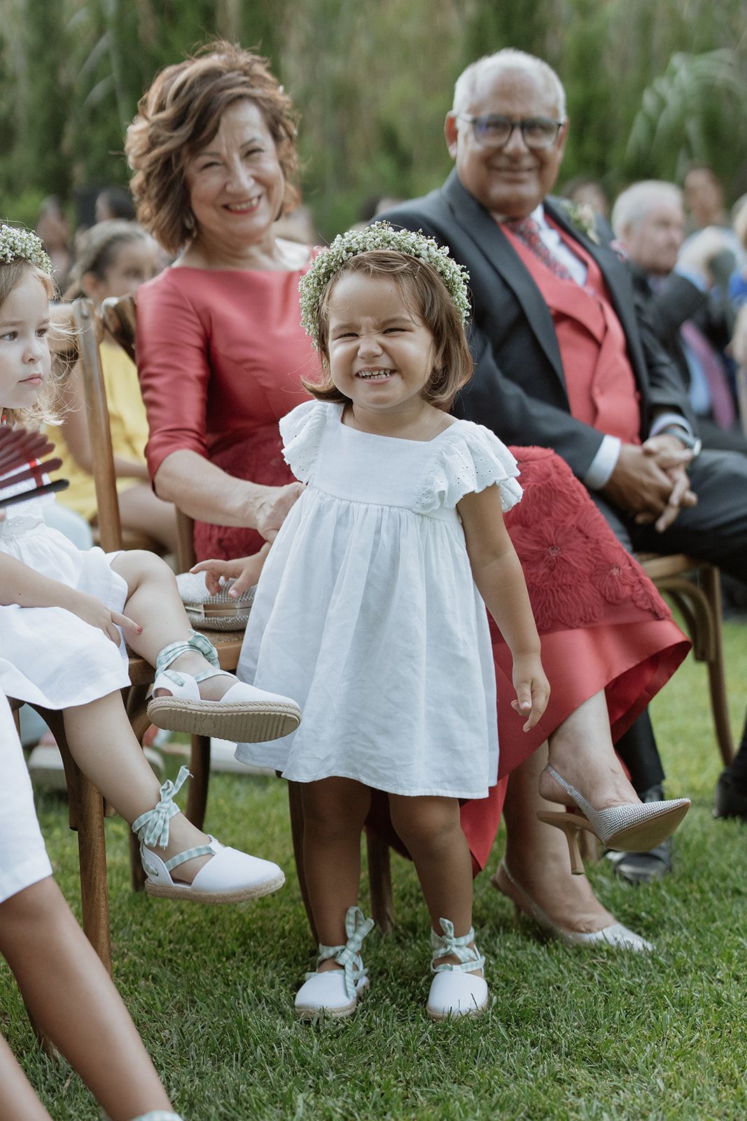 A little girl in a white dress stands among wedding guests on a grassy lawn.