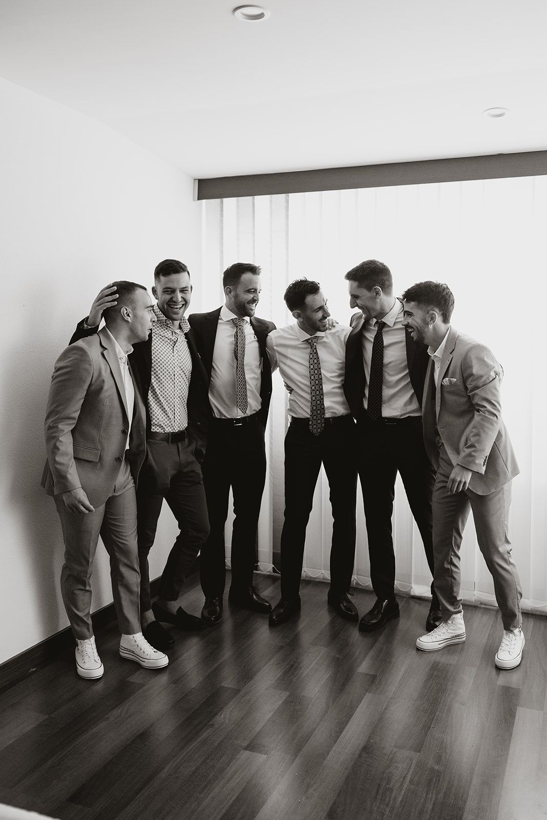 A group of groomsmen laugh together in formal wear during wedding preparations.