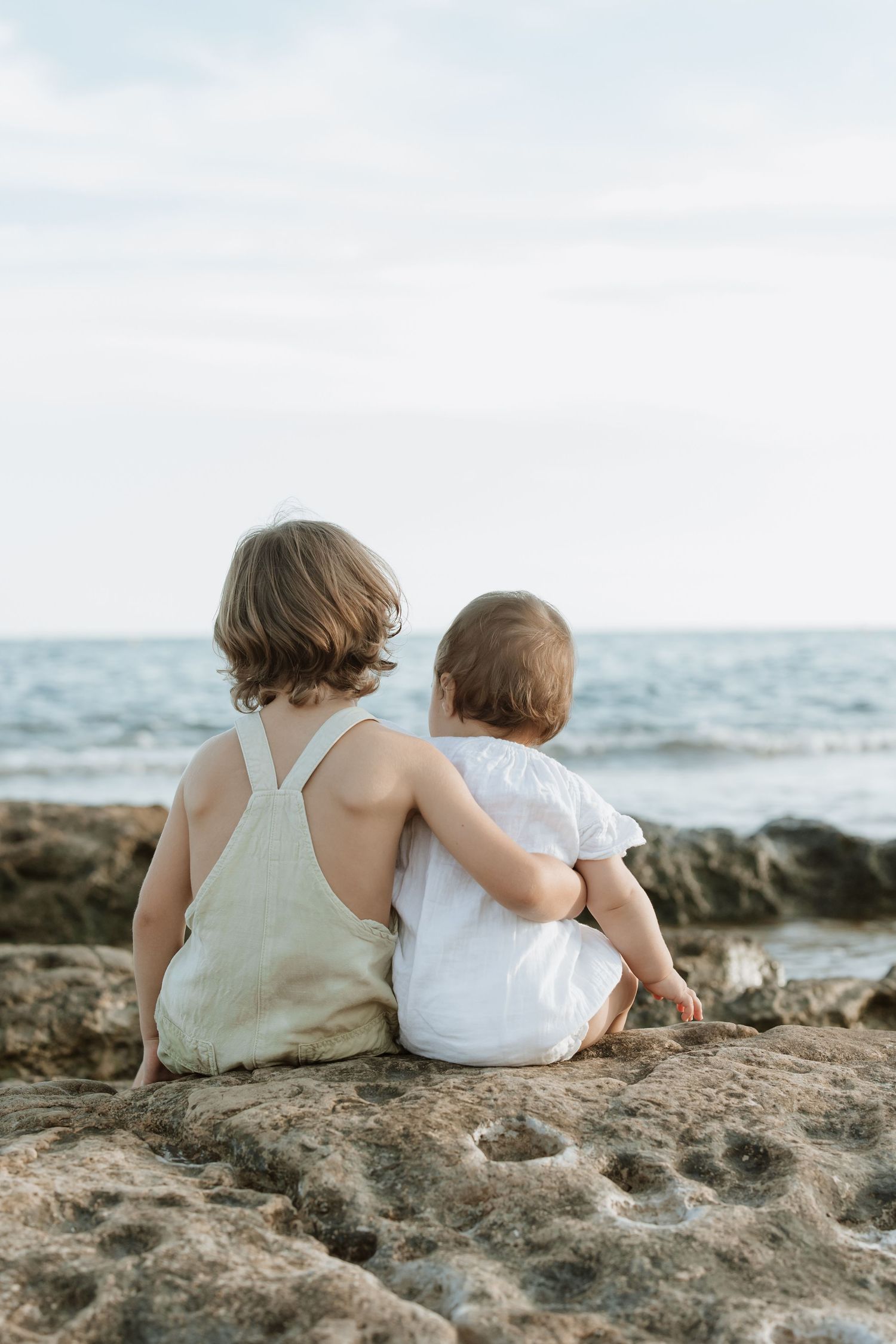 Two figures sit together on a rocky beach overlook facing the ocean horizon.