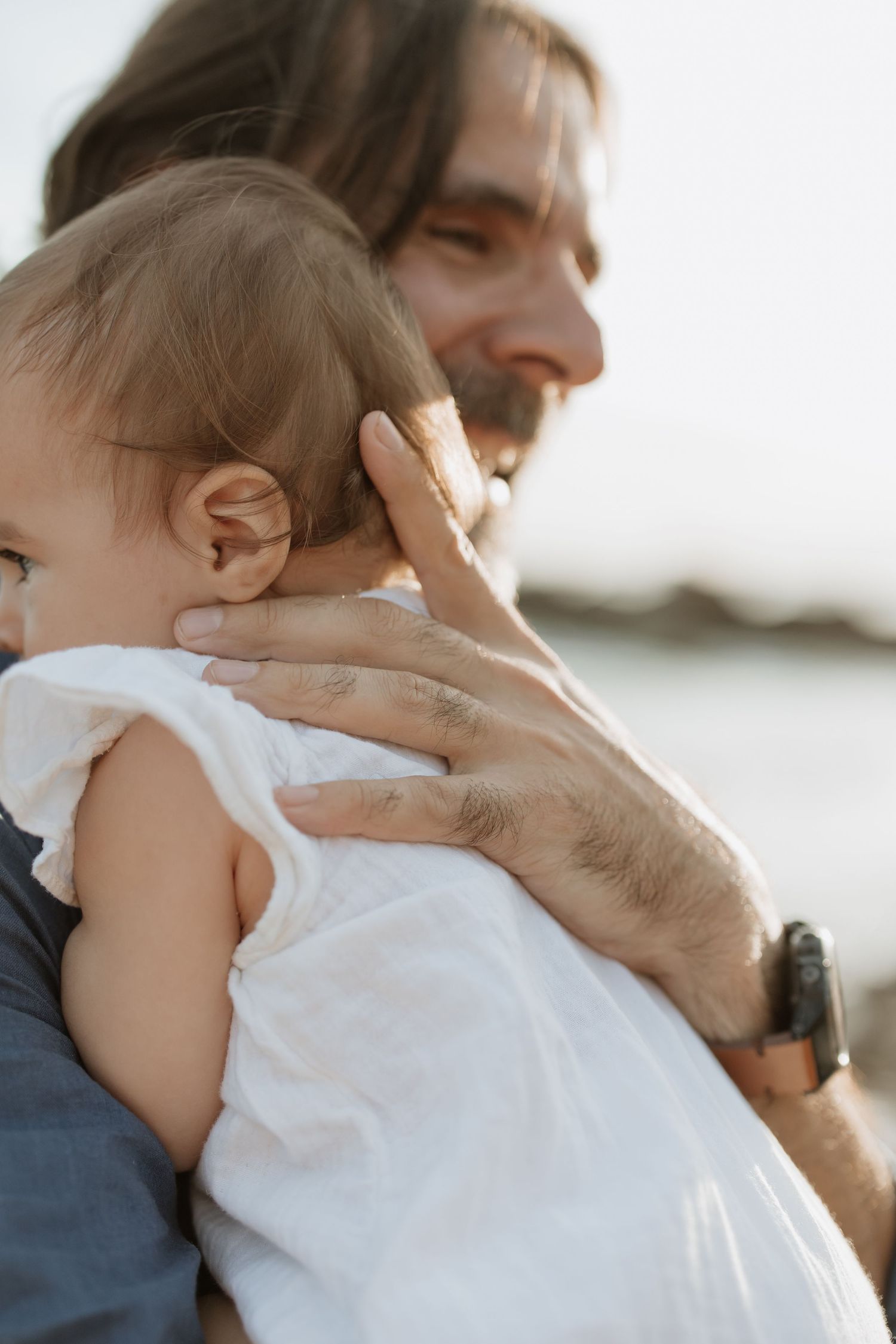Intimate moment between adult and baby in white dress against bright sky.
