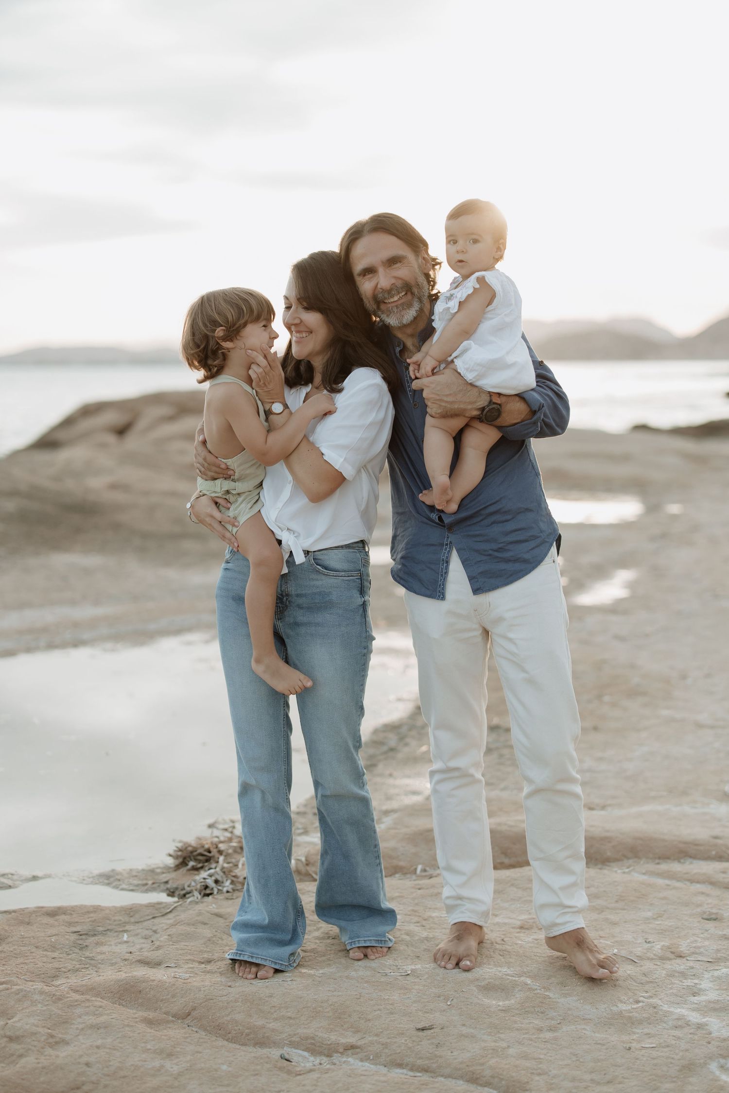 A family of four shares a tender moment together on a rocky beach at sunset.