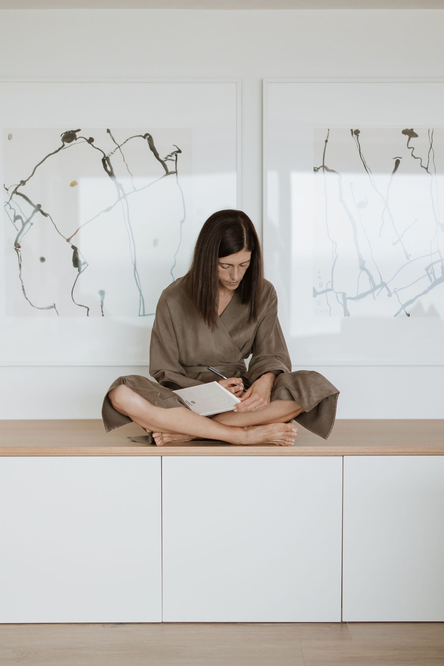Person in brown dress sitting cross-legged on white cabinet beneath minimal wall art.