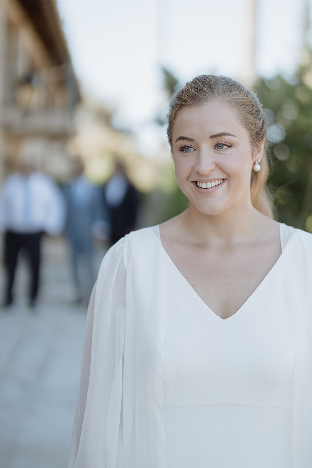 Bride wearing elegant white v-neck wedding dress with flowing sleeves smiles in outdoor setting.
