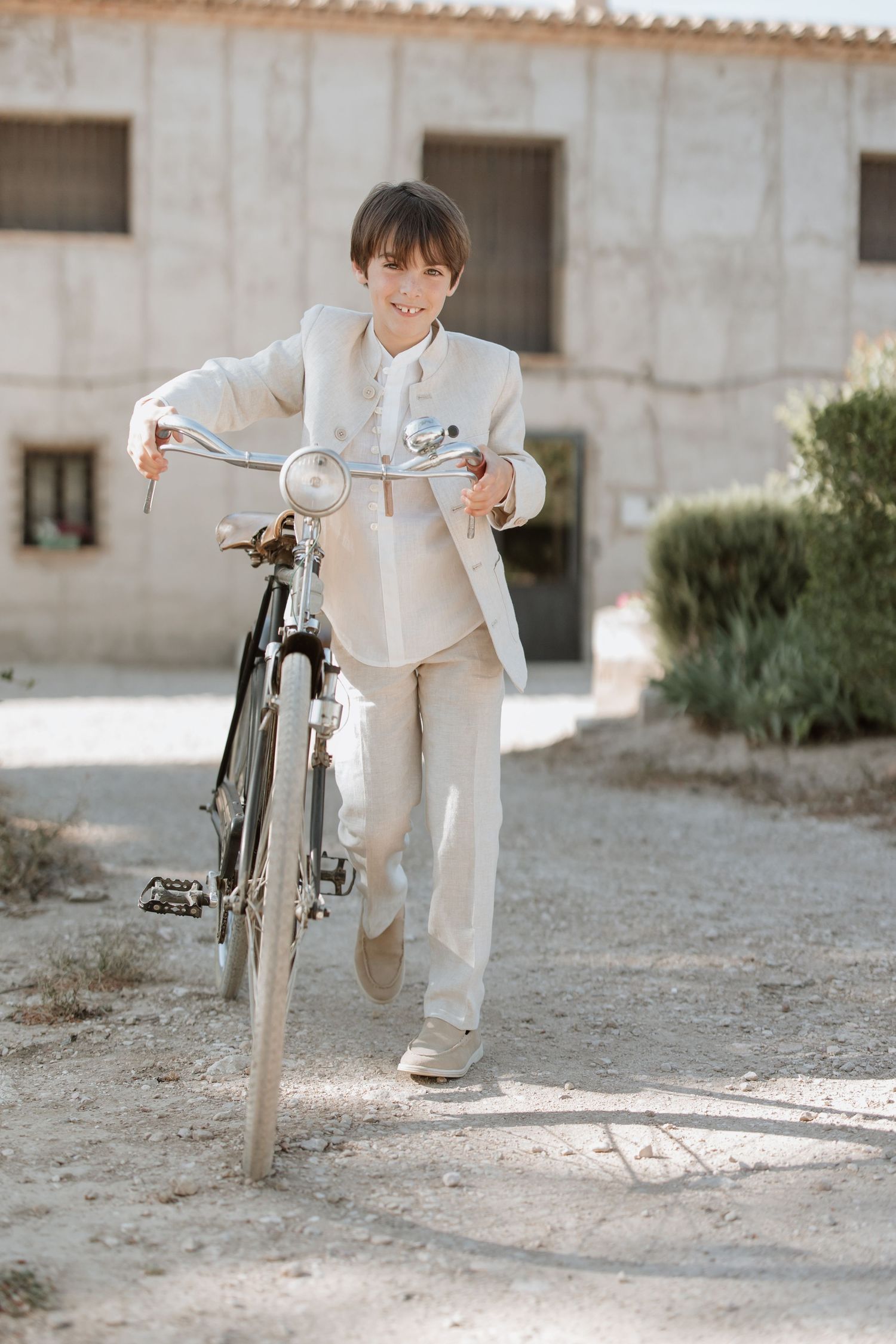 Person in light colored clothing standing next to a vintage bicycle on a sunlit gravel path near a stucco building.