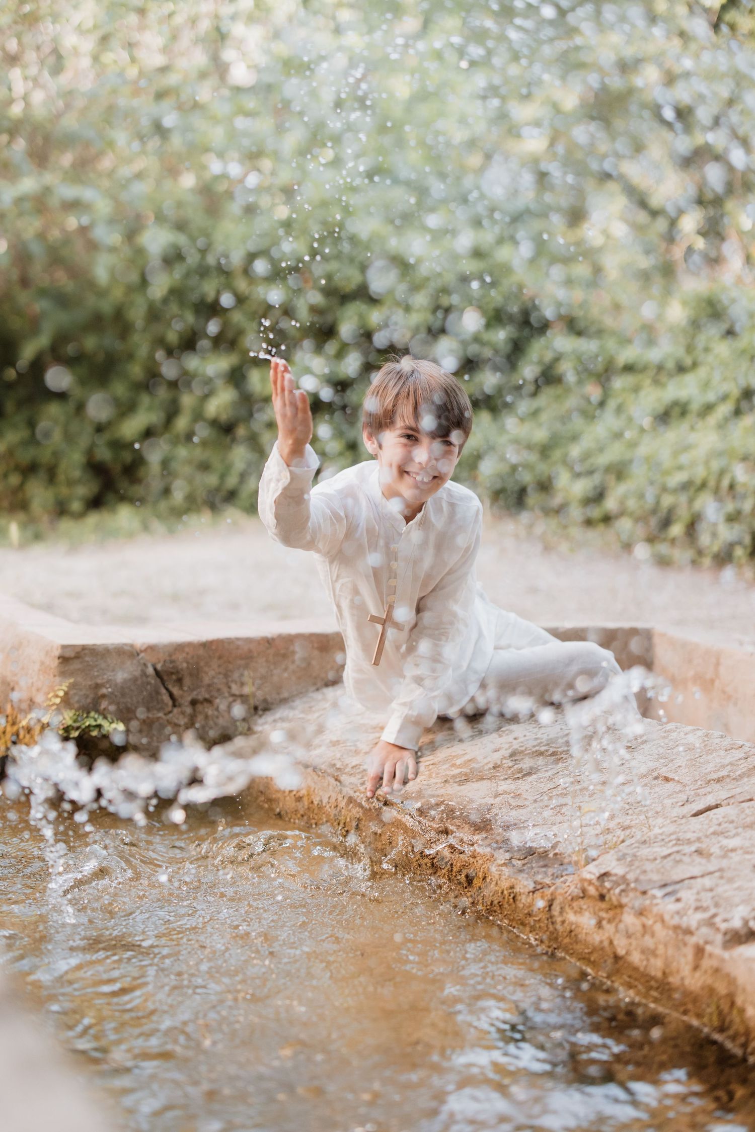 A person in white clothing sits by a stream splashing water on a sunny day amidst dreamy bokeh and nature.