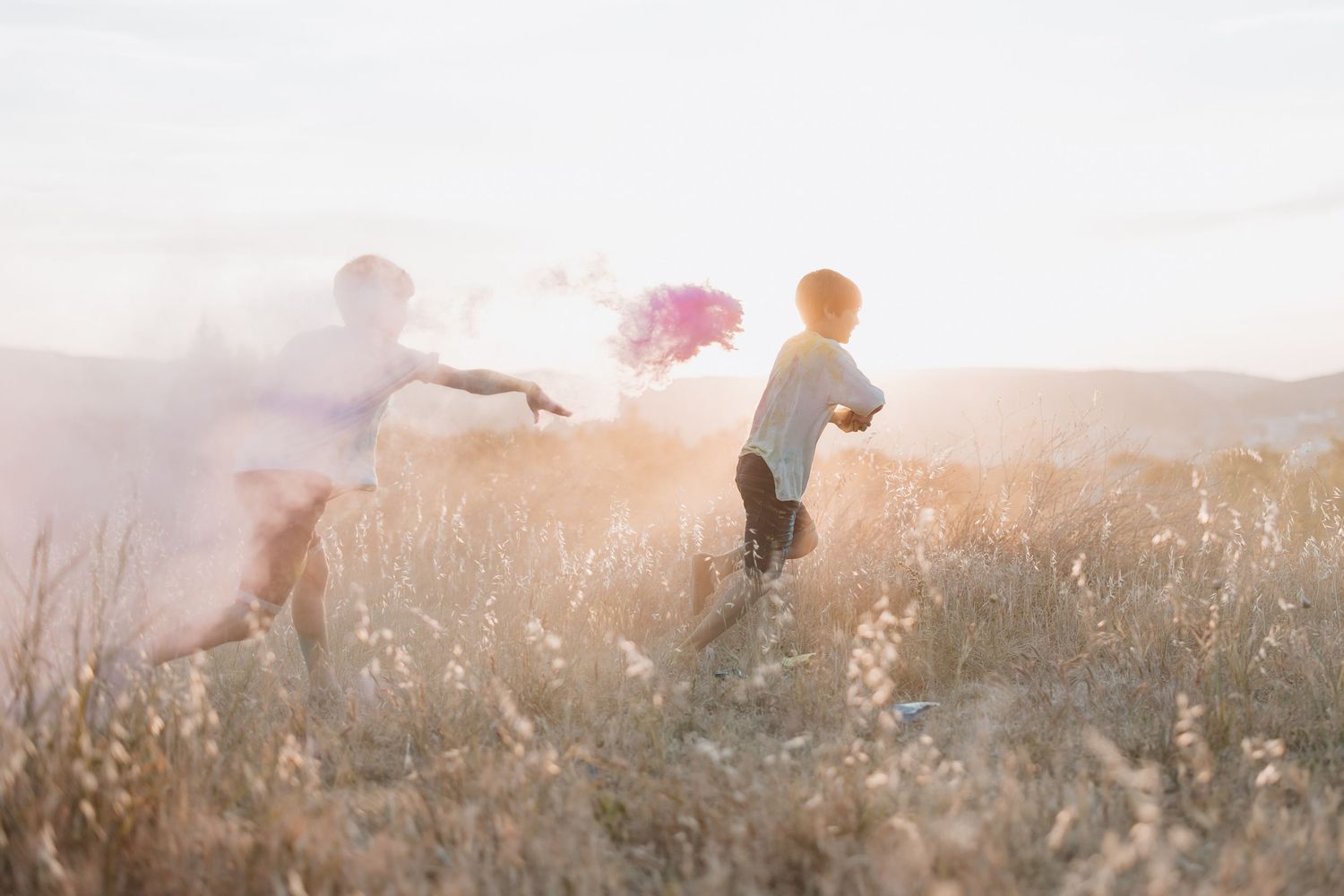 Silhouettes of people running through a hazy golden field at sunset with colorful smoke bombs creating a dreamy atmosphere.