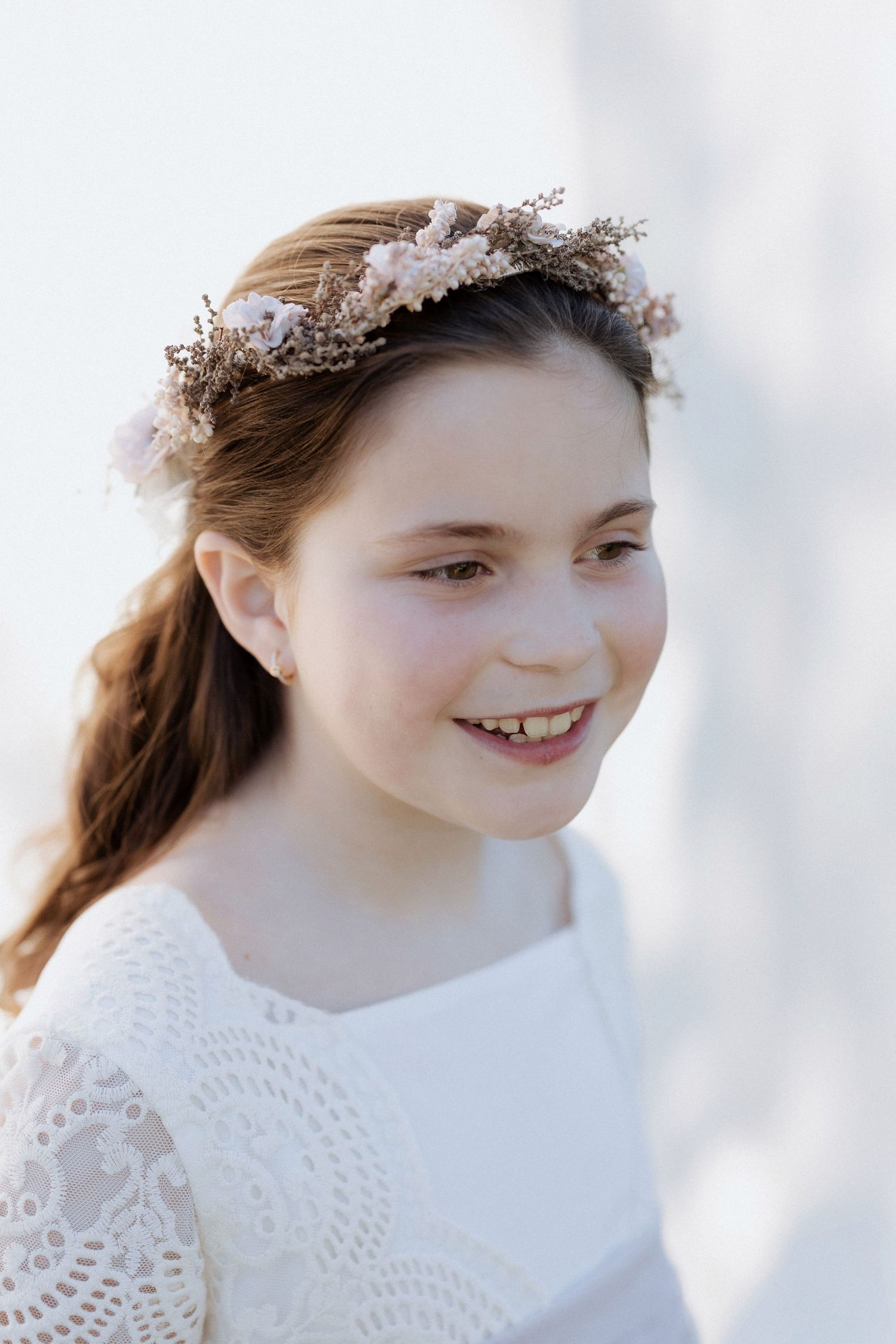 Smiling young girl wearing a floral crown and white lace dress, perfect for a First Communion or special occasion.