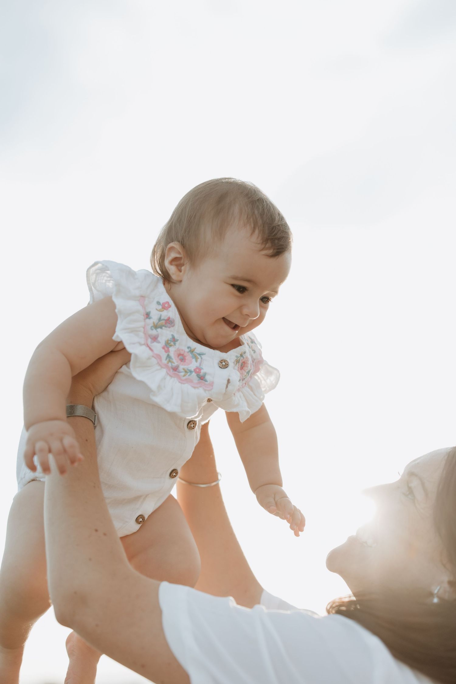 A baby in a white ruffled romper being lifted against a bright sky.