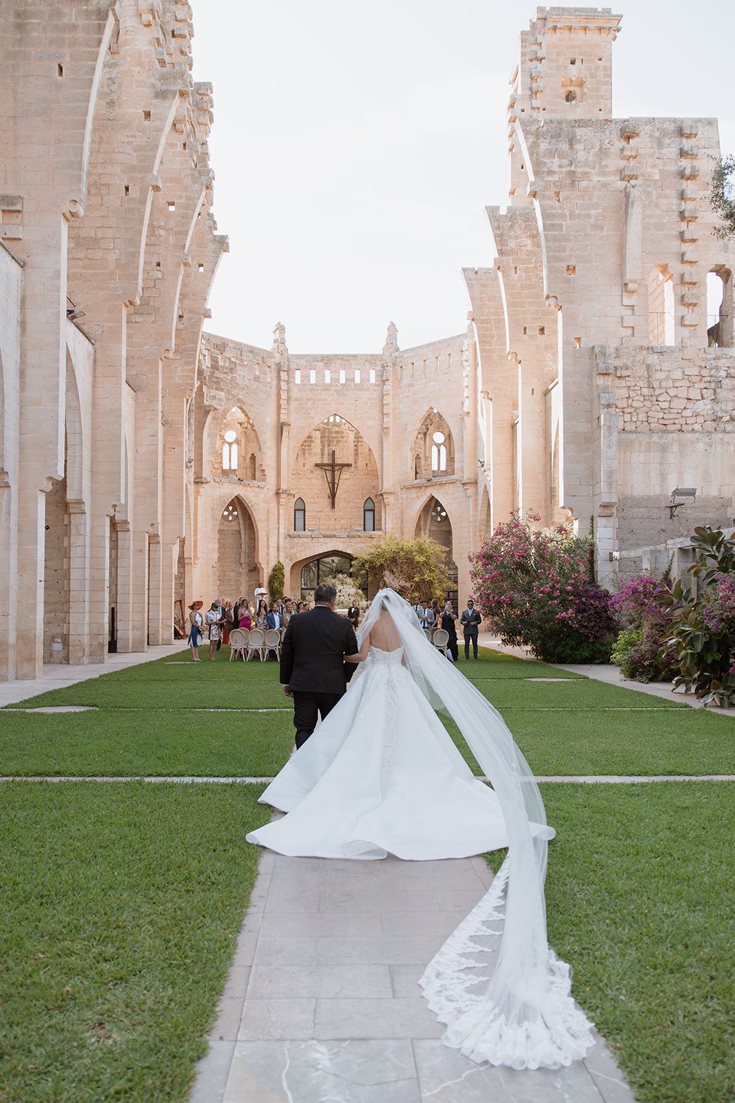 A wedding couple walks down a stone pathway towards a majestic gothic church with towering arches.