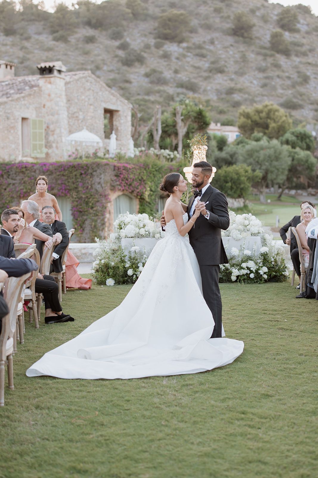 A romantic wedding ceremony takes place on a lush lawn with a stone villa backdrop and floral arrangements.