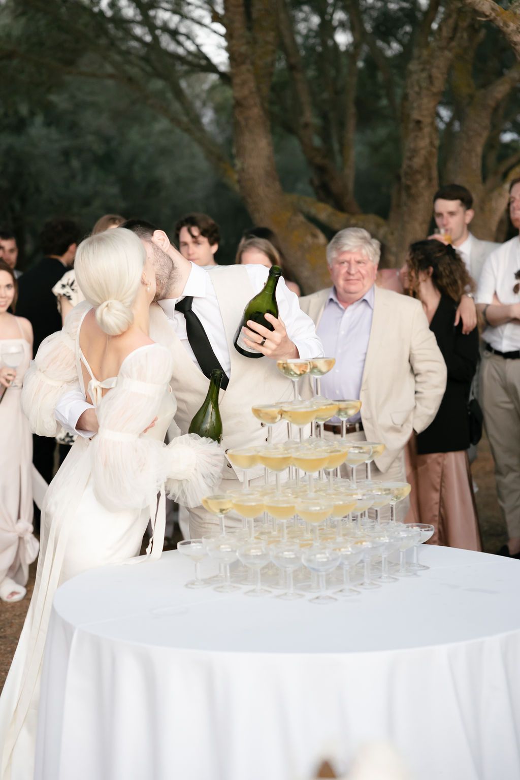 Guests gathering around a champagne tower at an outdoor wedding celebration.