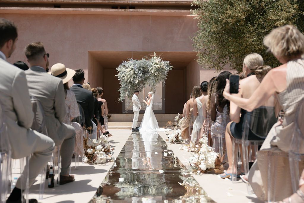 Wedding ceremony with mirrored aisle runner and guests seated on both sides under sunny skies.