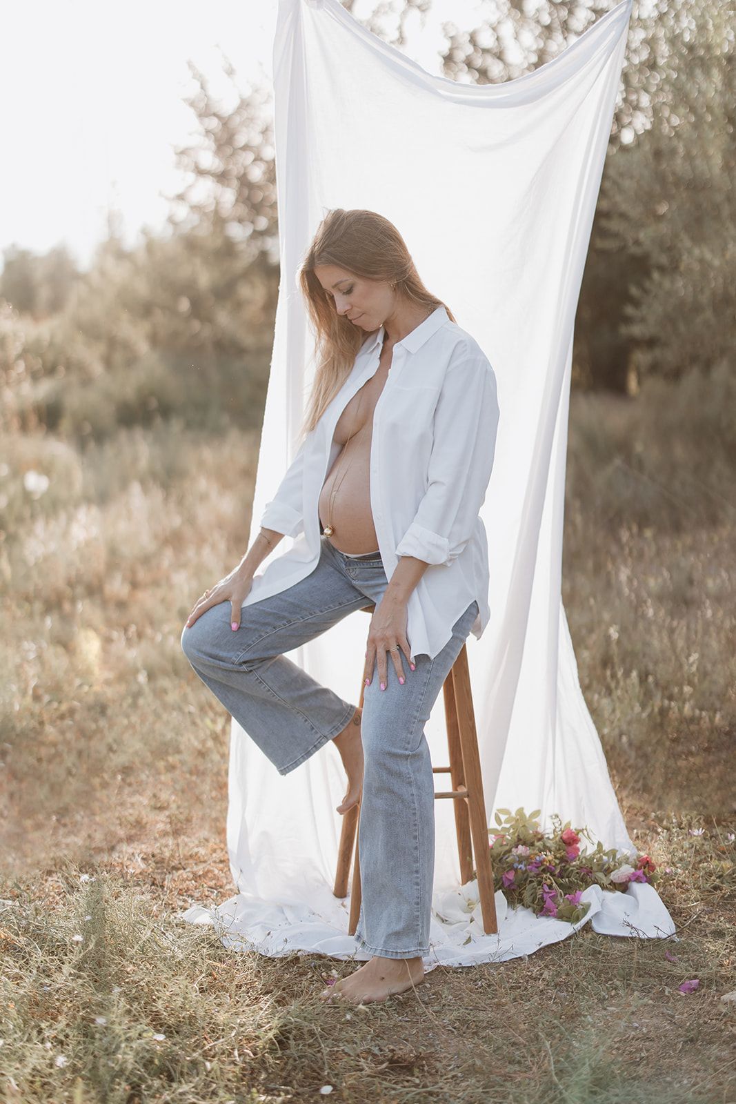 Casual maternity portrait featuring white button-down shirt and jeans against natural backdrop.