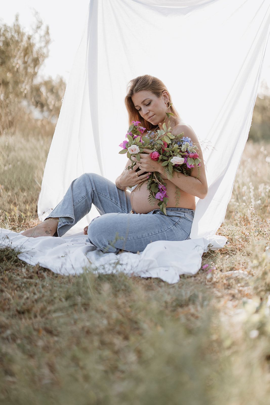 A maternity photoshoot featuring a casual look with jeans and floral bouquet against a white backdrop outdoors.