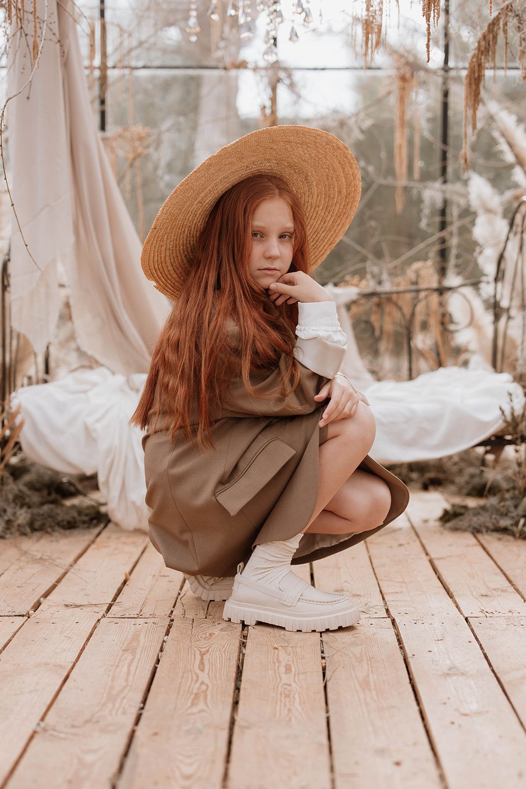 Whimsical greenhouse portrait with straw hat, brown coat and white boots.