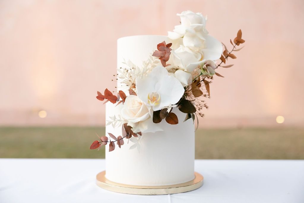 Elegant white wedding cake decorated with fresh orchids and copper-colored foliage on a gold base.