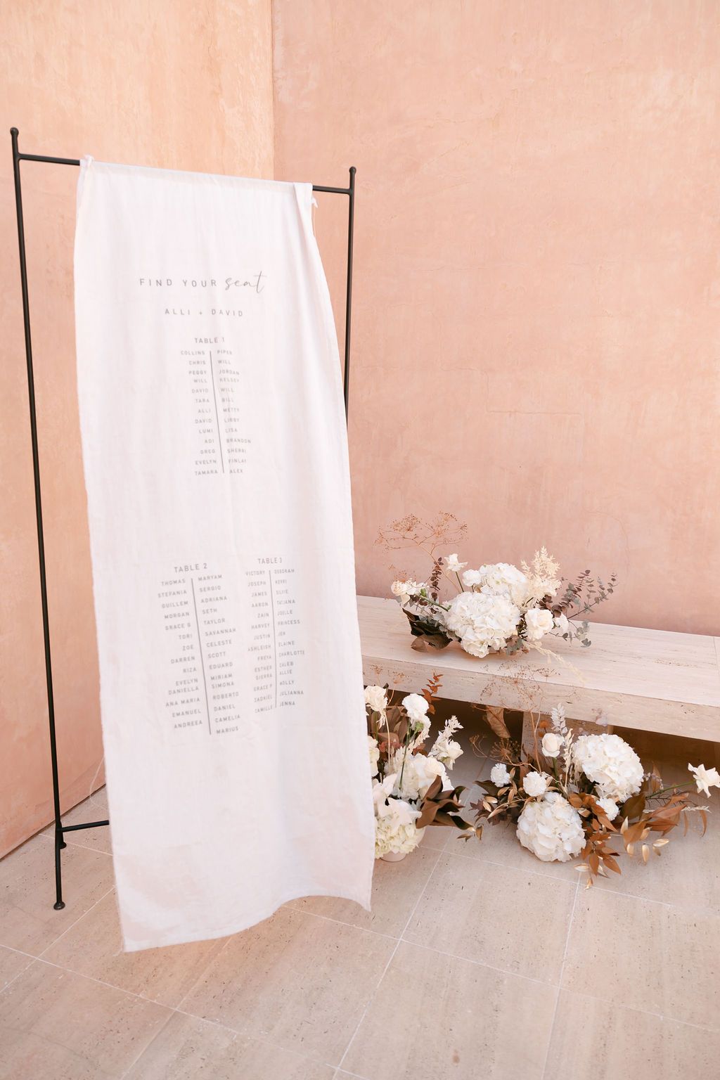 White seating chart display on a stand with delicate floral arrangements on a pink wall setting.