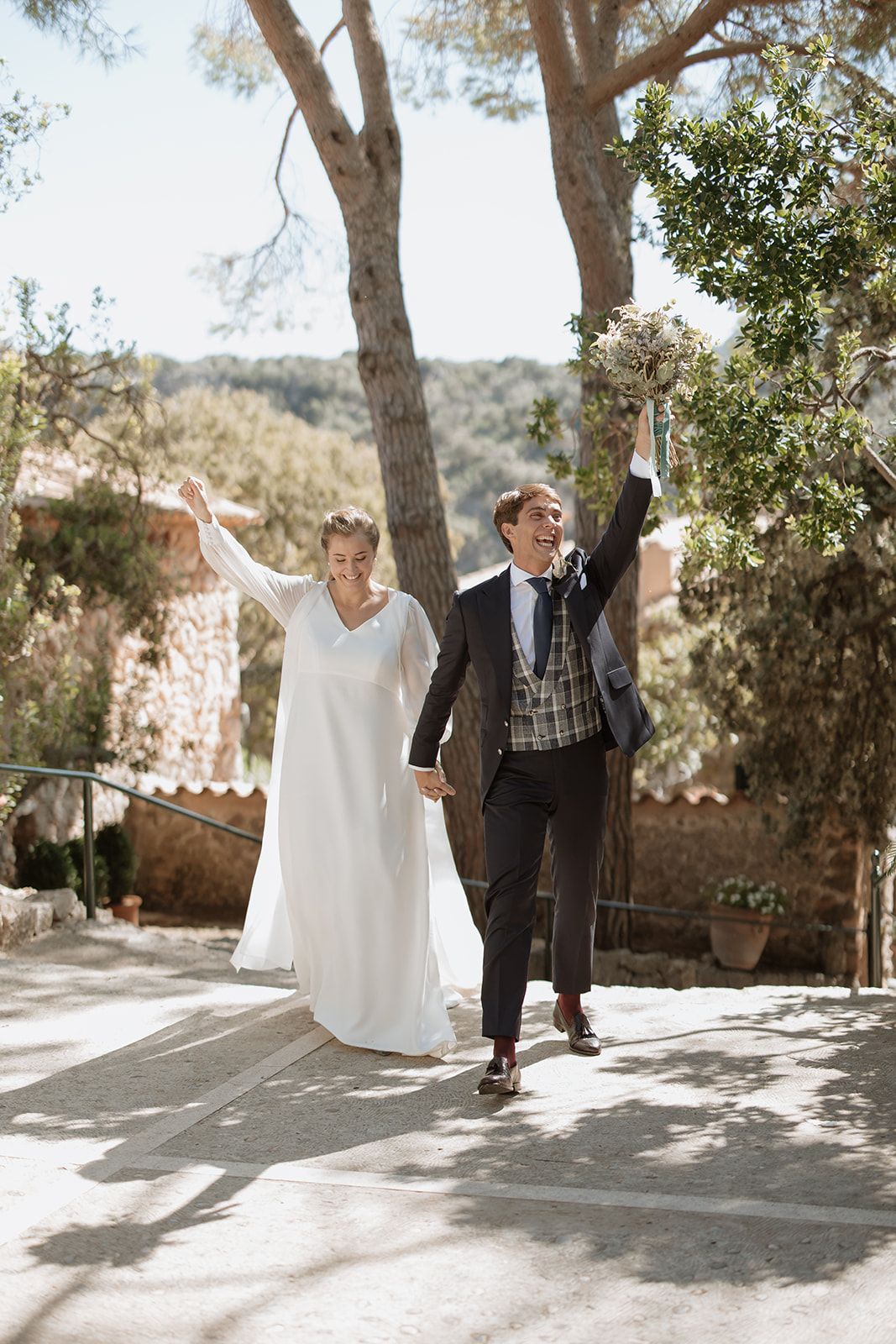 Bride and groom dance playfully in sunlit outdoor venue surrounded by trees.
