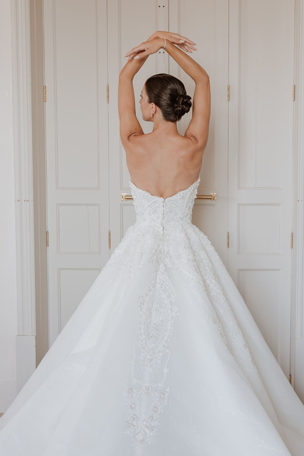 Artistic back view of bride in strapless ball gown with raised arms against white doors.