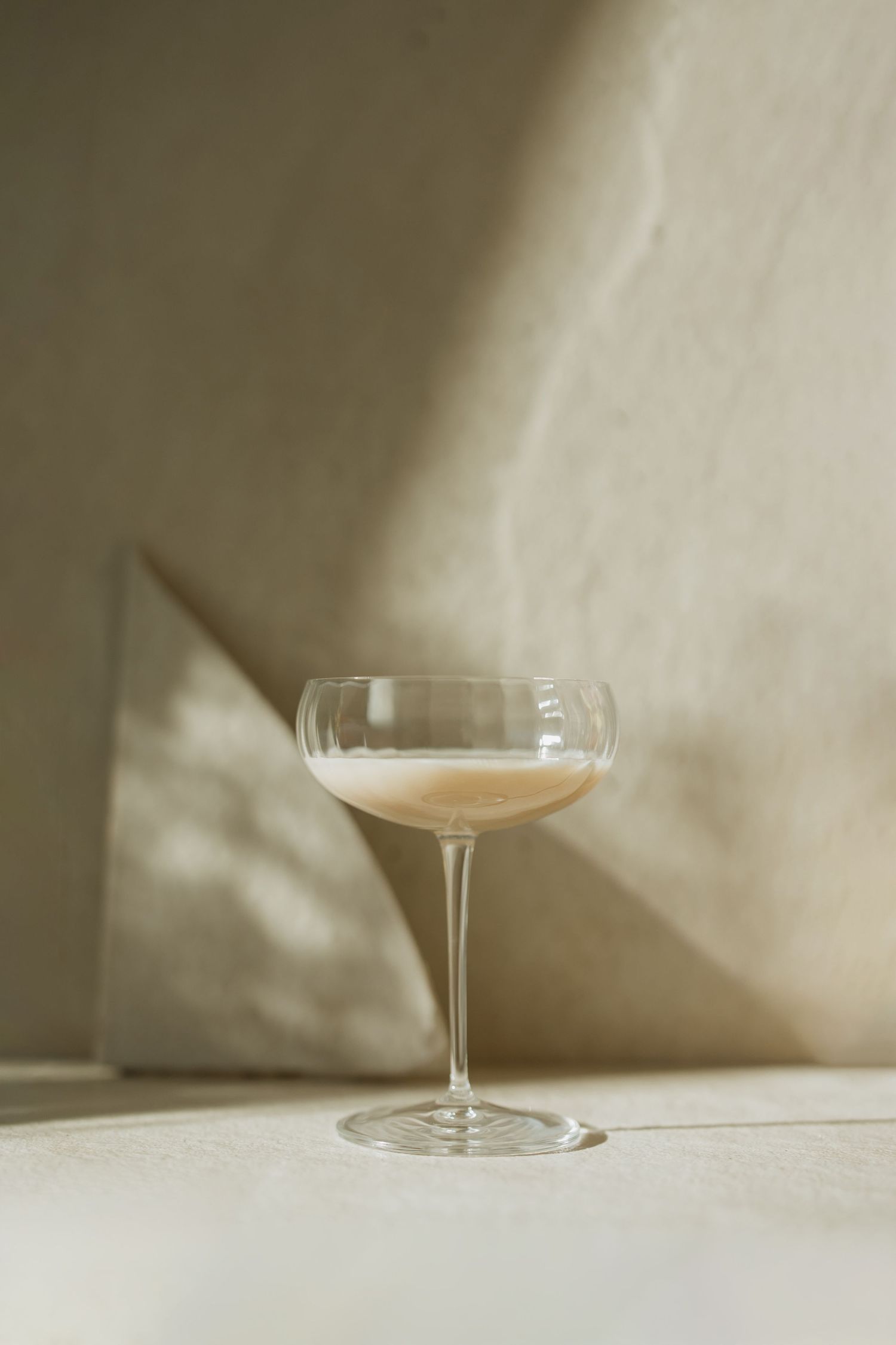 An elegant coupe glass filled with a pale cocktail casting shadows on a minimalist white surface.
