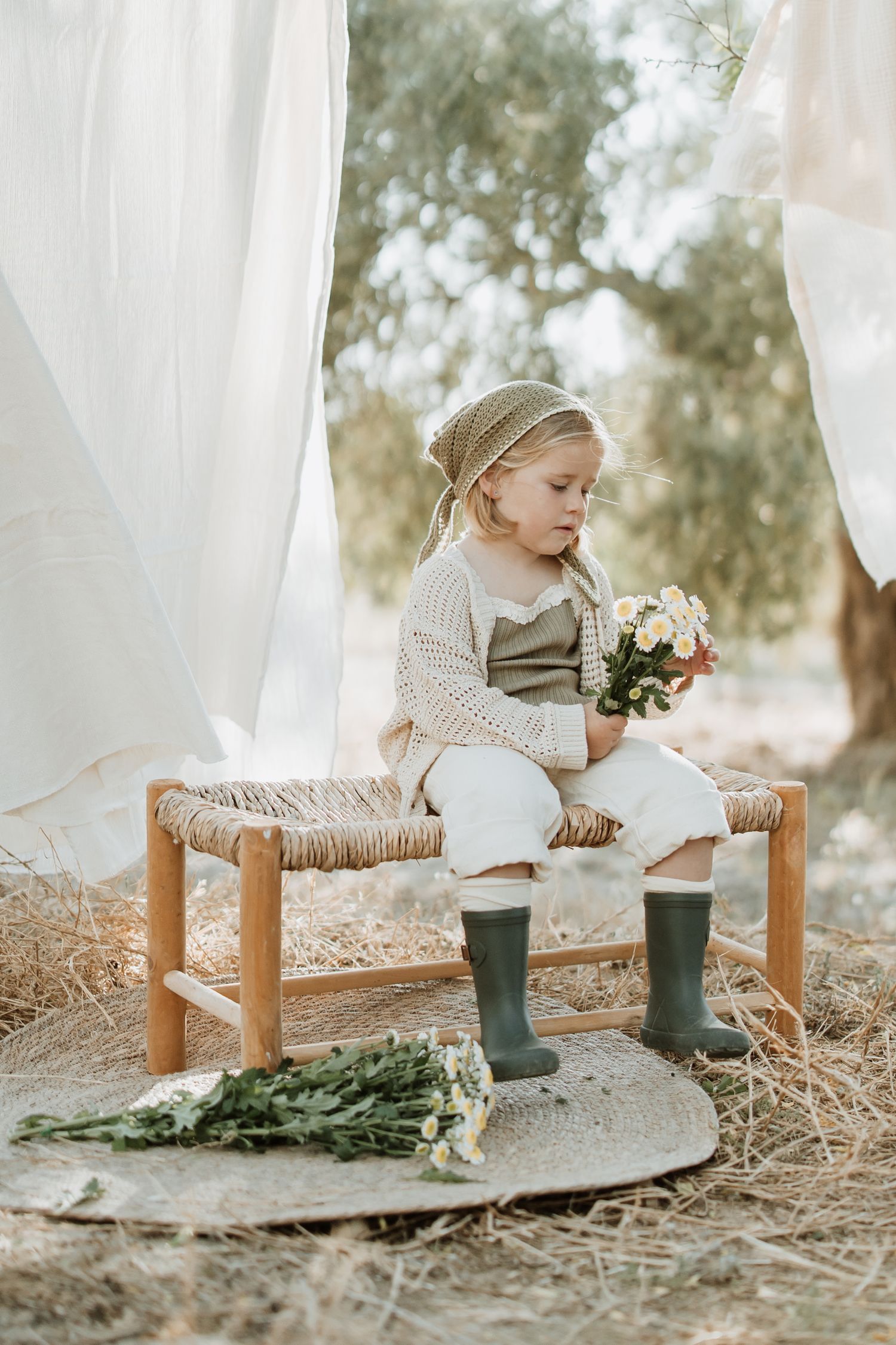Child wearing headscarf and knit sweater holding flowers on rustic bench in outdoor setting.