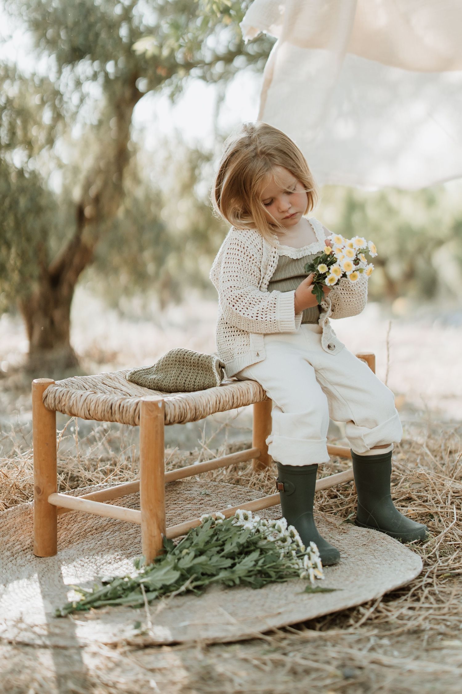 Child in white outfit and boots sits on rustic stool examining flowers in natural outdoor setting.