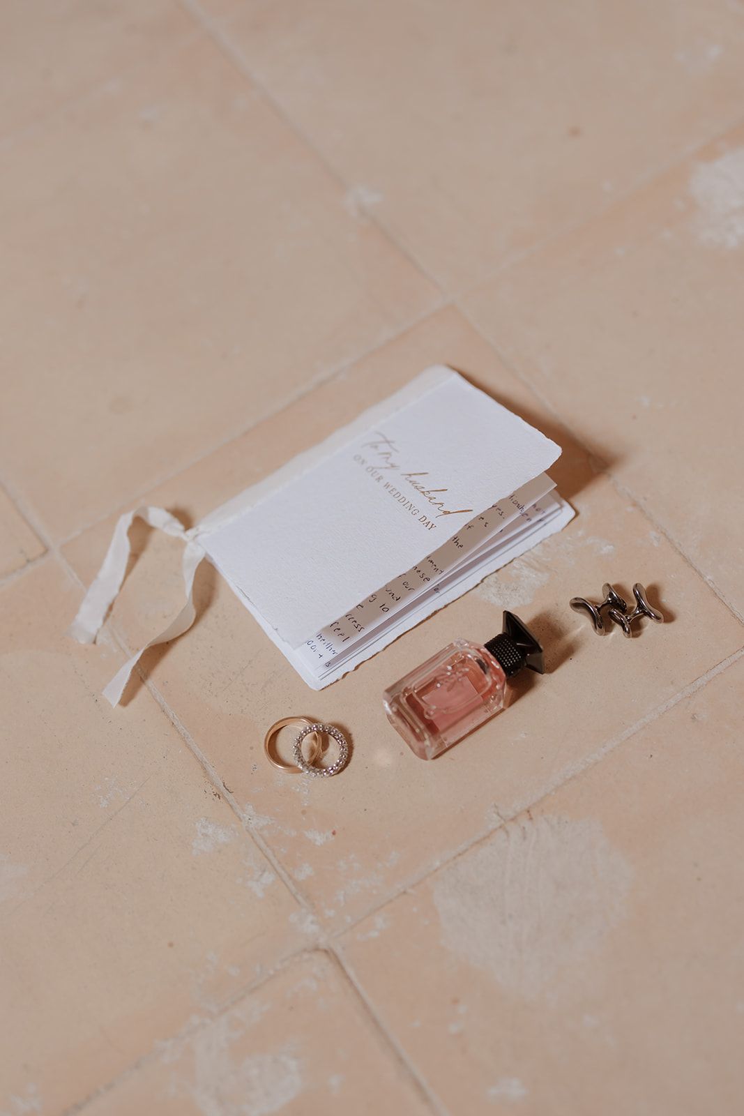 Wedding details flatlay with rings, perfume bottle and invitation on beige tile floor.