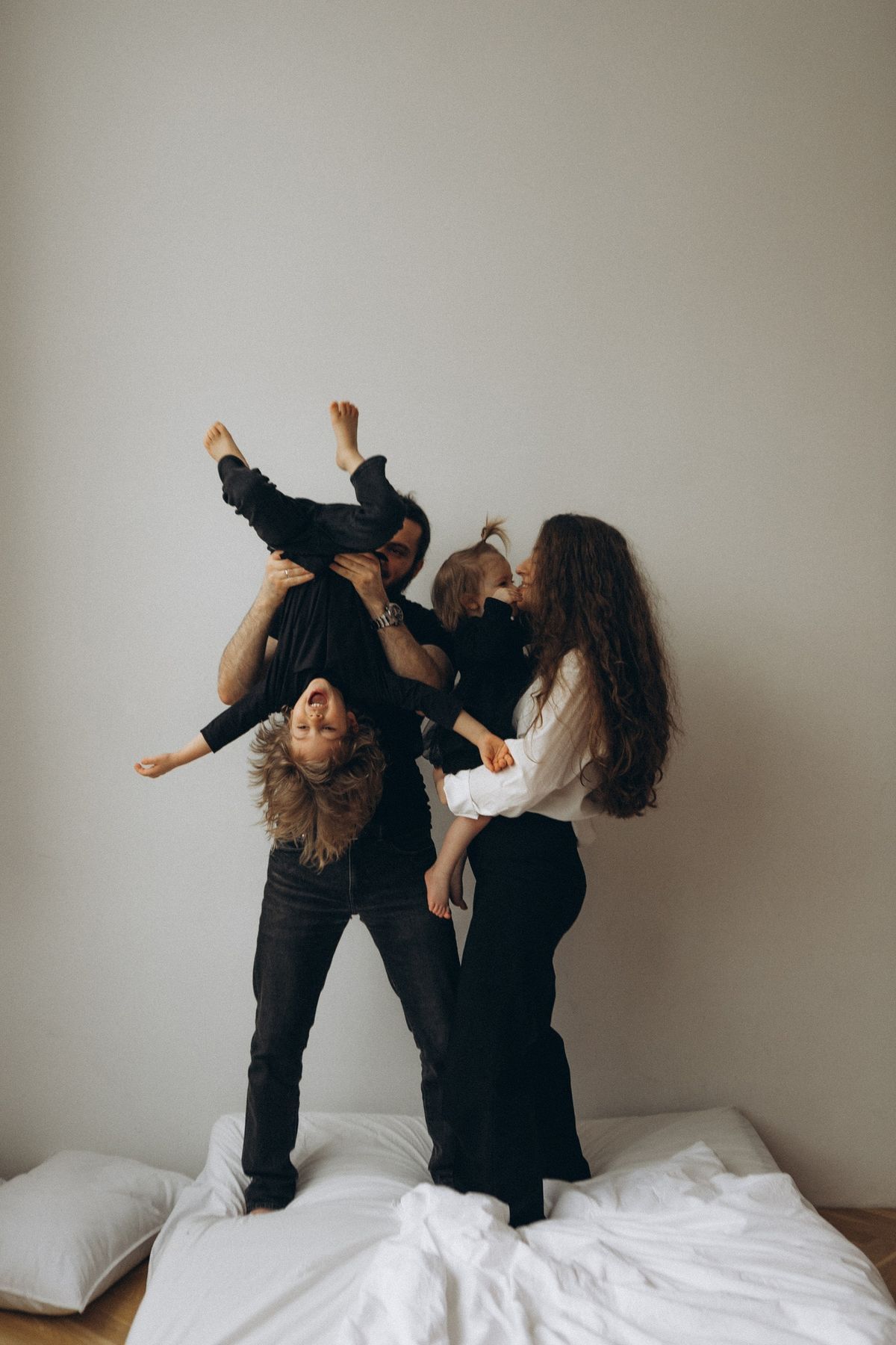 Two people in black outfits playfully jumping and dancing on a white bed against a neutral wall.
