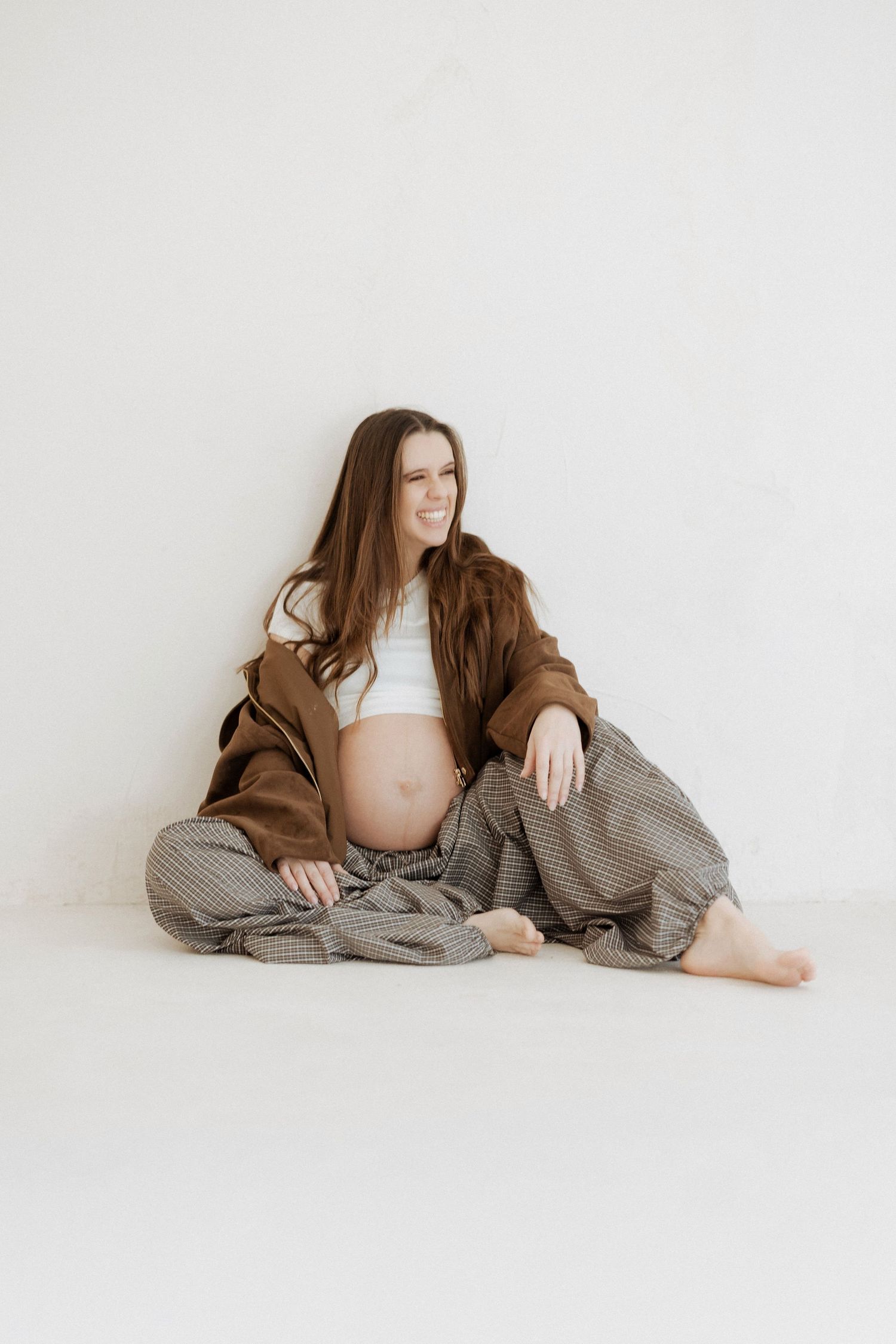 A figure in a white crop top and oversized brown cardigan reclines against a white backdrop in a minimalist fashion pose.