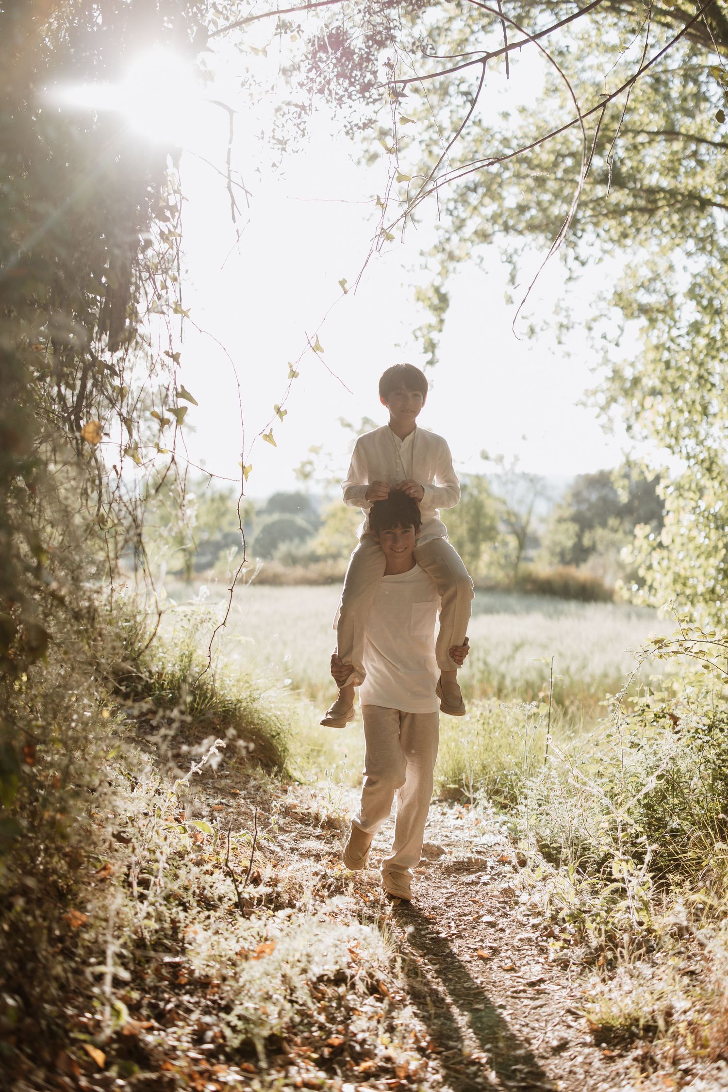 Silhouetted figures walking on a sun-dappled nature path through trees and tall grass in dreamy golden light.