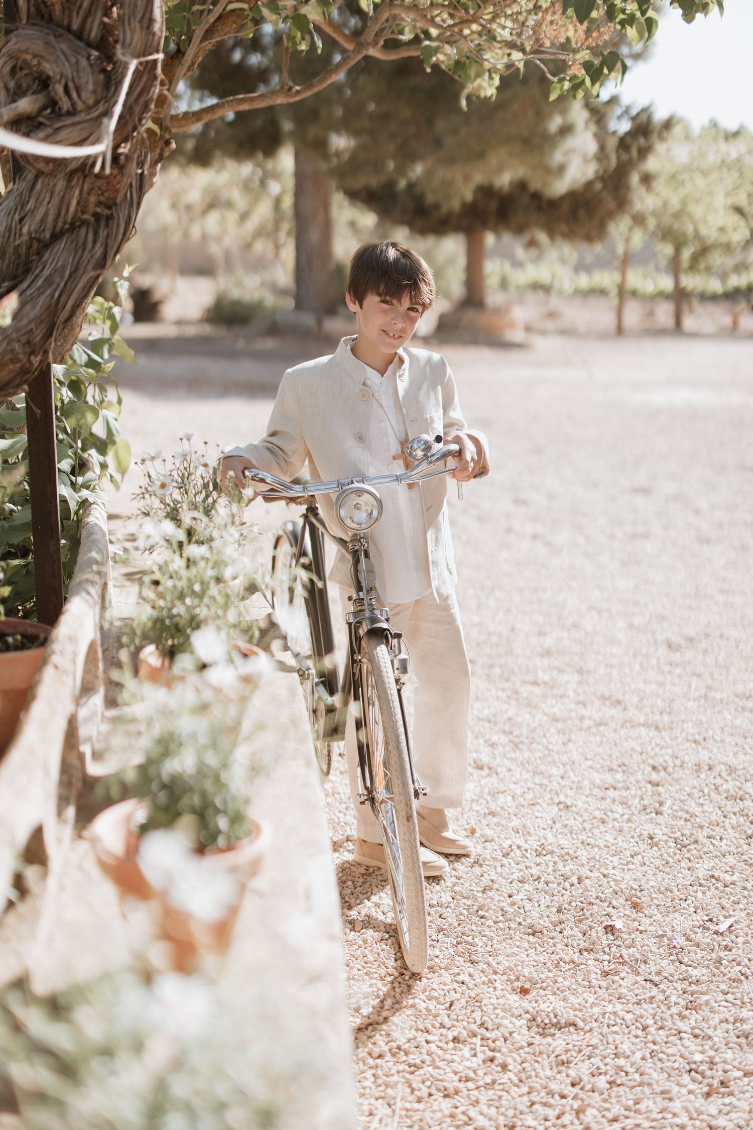 Someone in a light colored outfit leans on a vintage bicycle on a gravel path surrounded by greenery and trees.