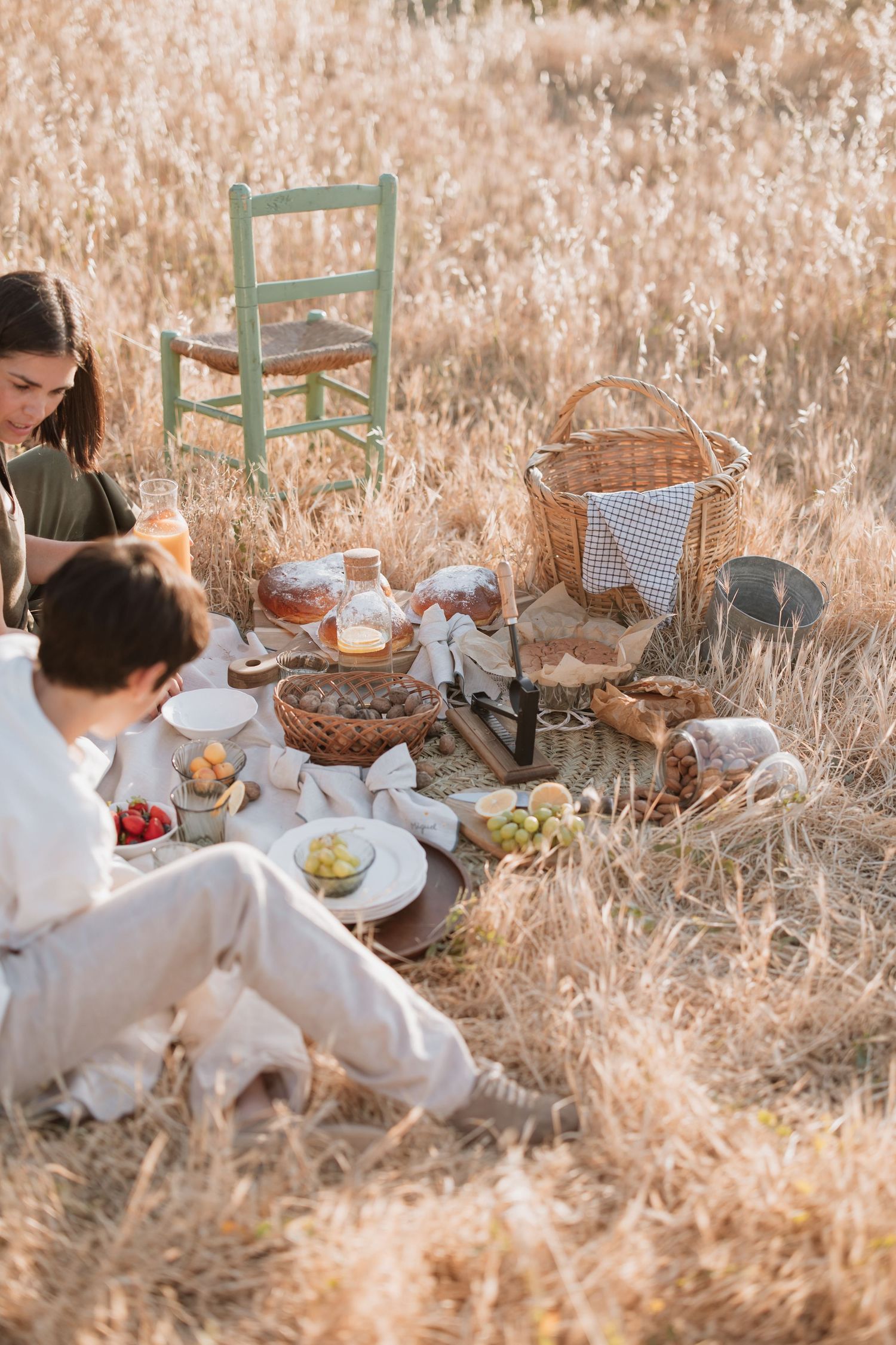 A cozy outdoor picnic scene with wicker baskets, blankets and rustic chairs set up in golden grass during sunset.