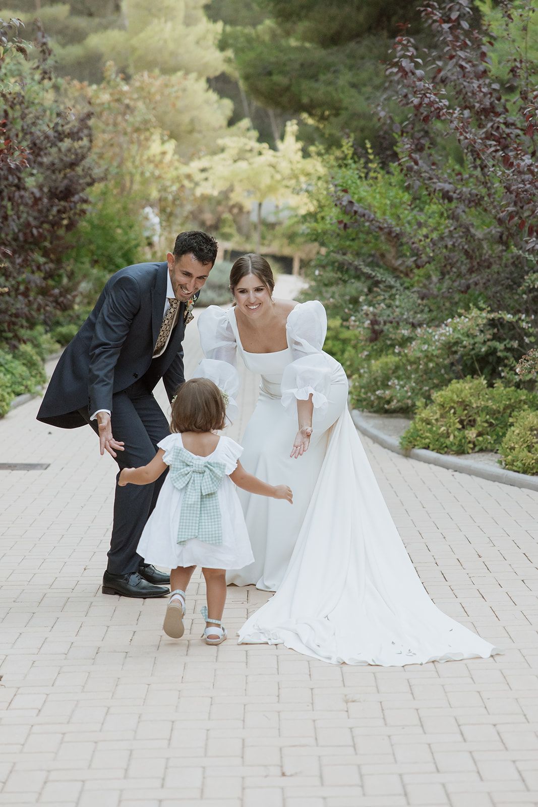 Wedding couple shares sweet moment with young flower girl on brick pathway.