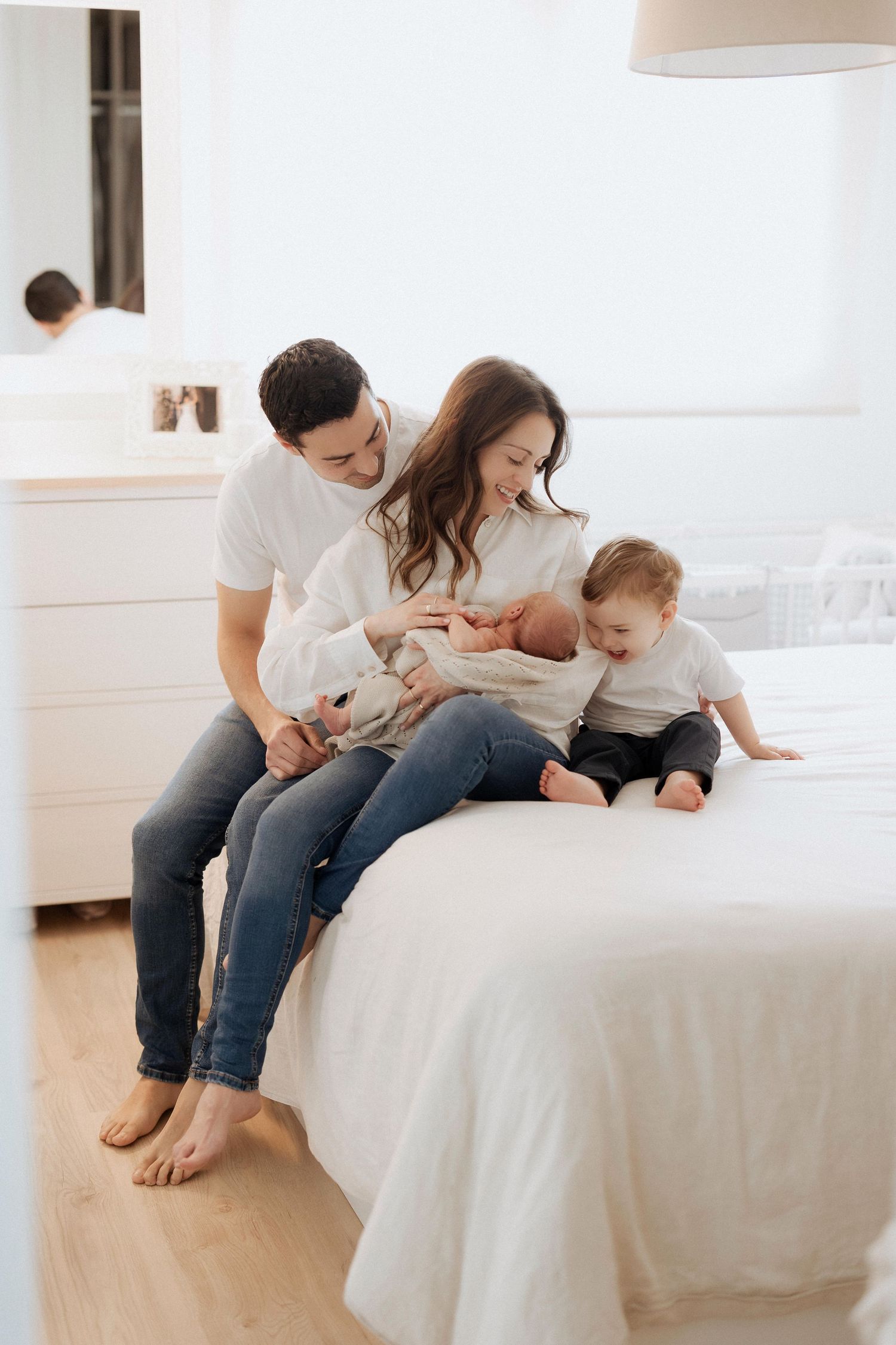 Family relaxing together on white bed wearing casual clothes in bright minimalist bedroom setting.