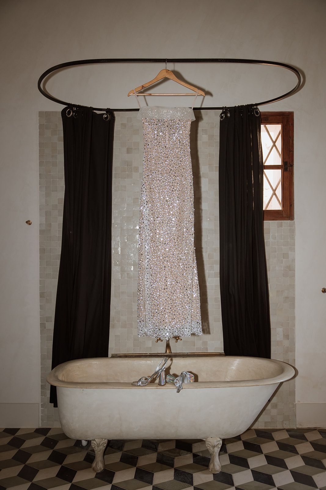 Sparkly wedding dress hanging above clawfoot bathtub with black curtains and checkered floor.