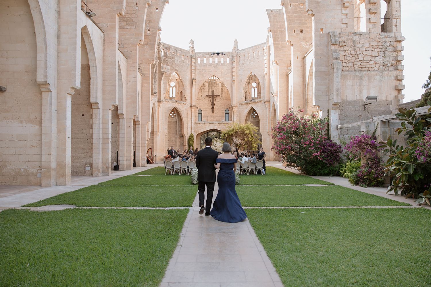 Couple walking through the courtyard of a gothic stone cathedral with arched architecture and manicured lawn.