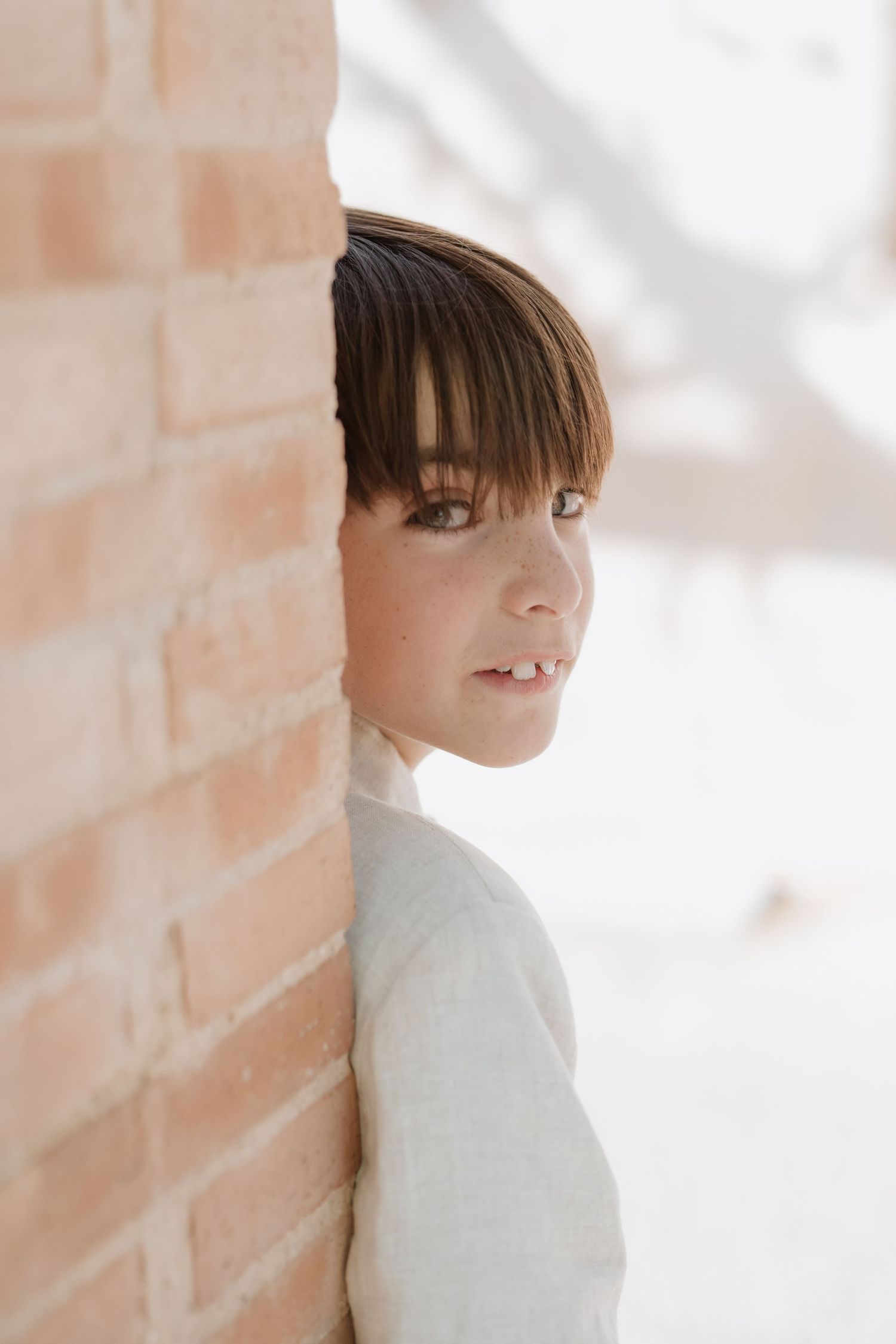 Portrait of someone peeking around a brick wall corner while wearing a white sweater in soft natural lighting.