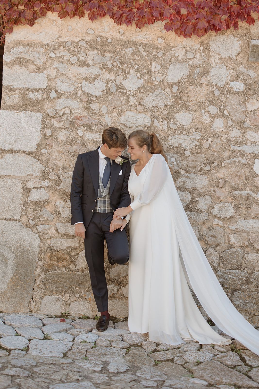 Newlyweds share tender moment against stone wall with autumn foliage.