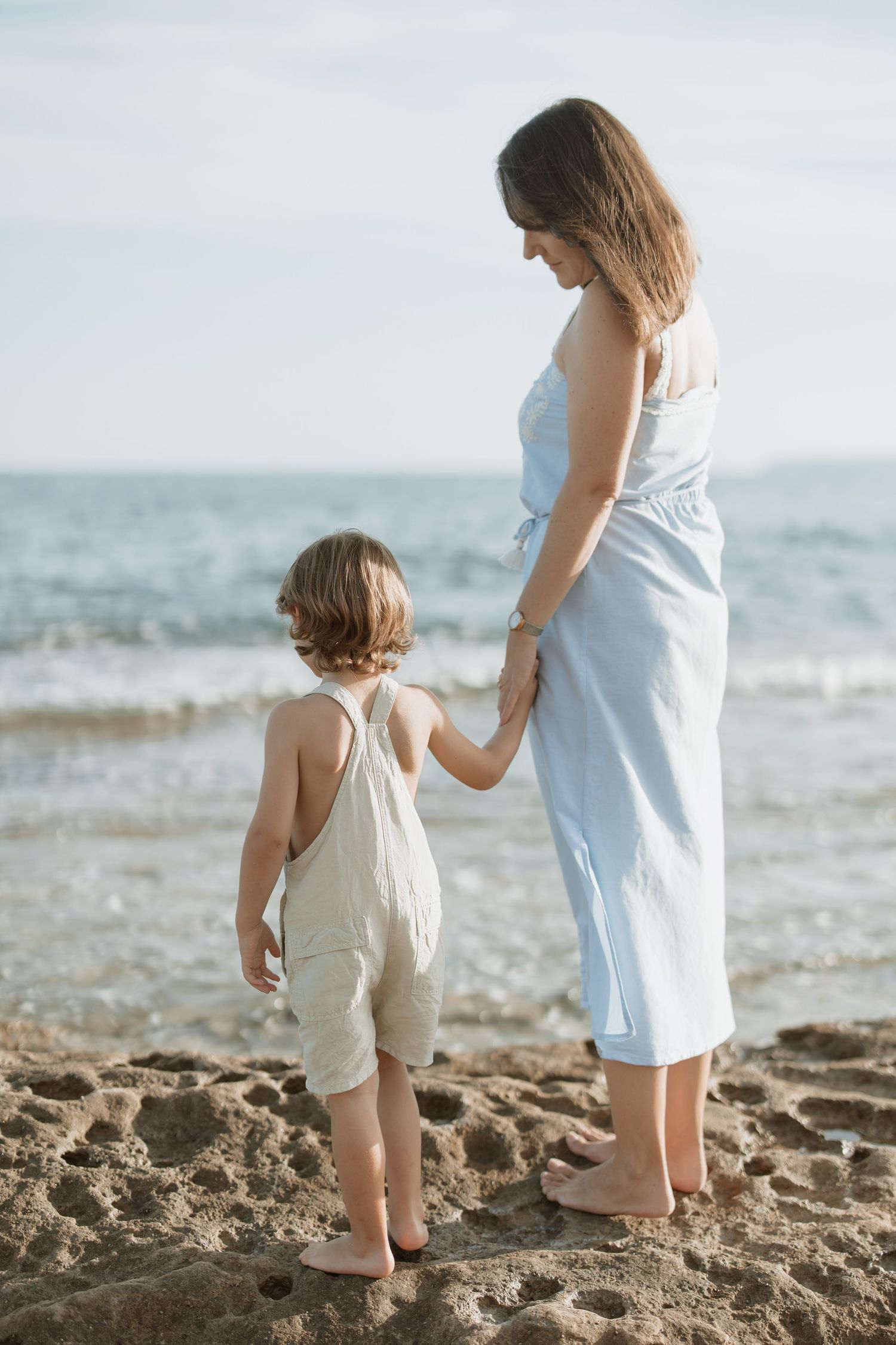 Mother and child holding hands while standing on sandy beach with ocean waves in background.