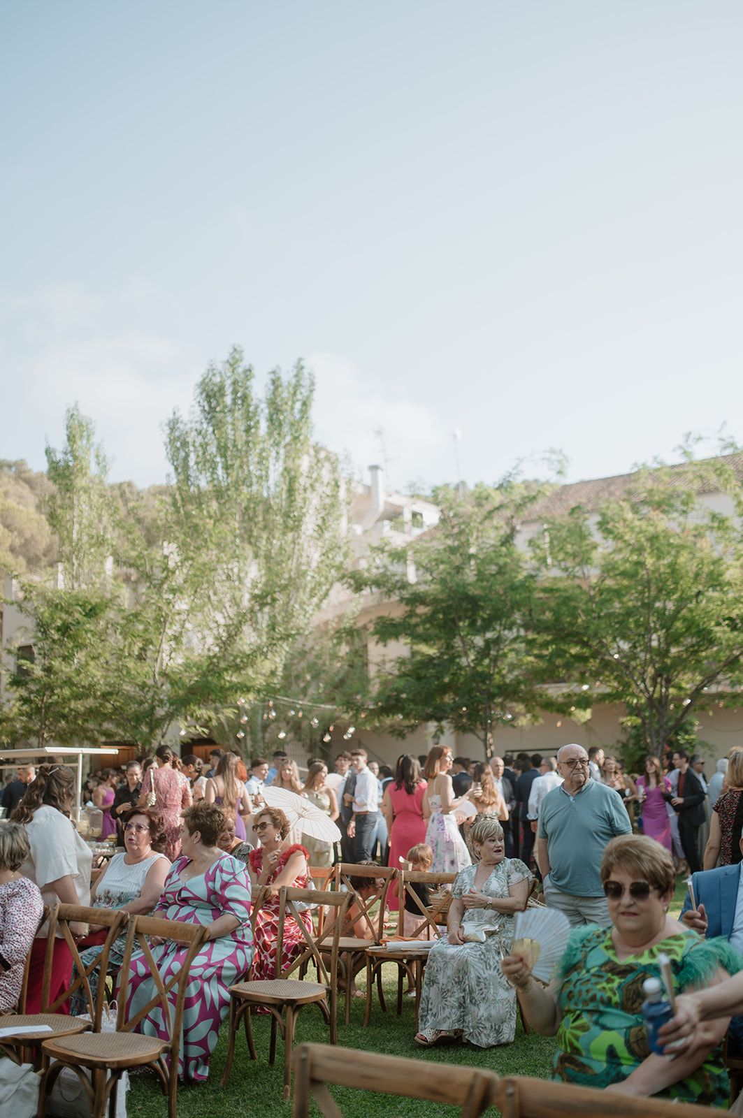 Wedding guests gather outdoors under trees with wooden chairs arranged for a casual garden ceremony.
