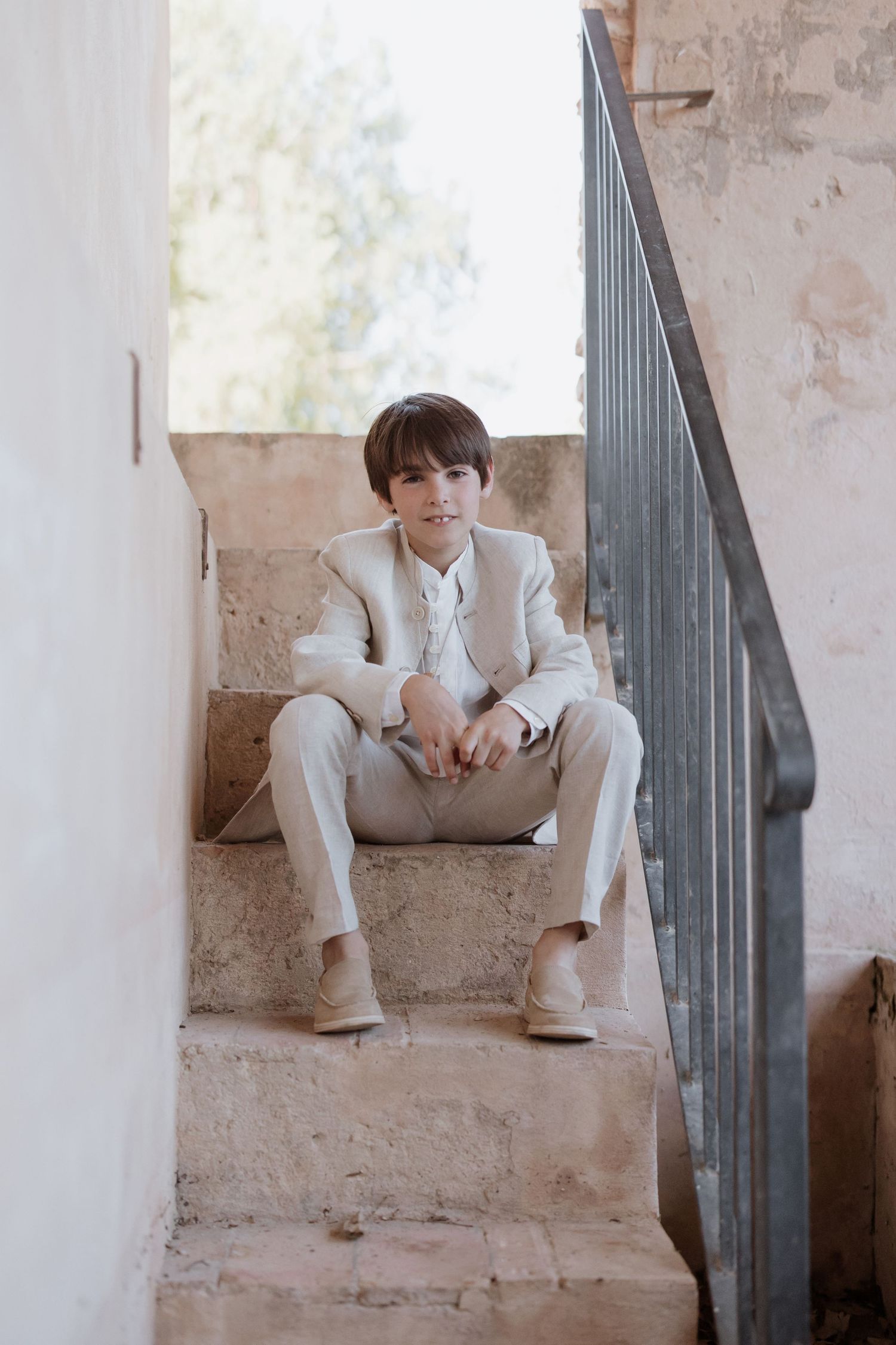 A person in a beige suit sits casually on concrete stairs against a light colored wall with a metal railing.