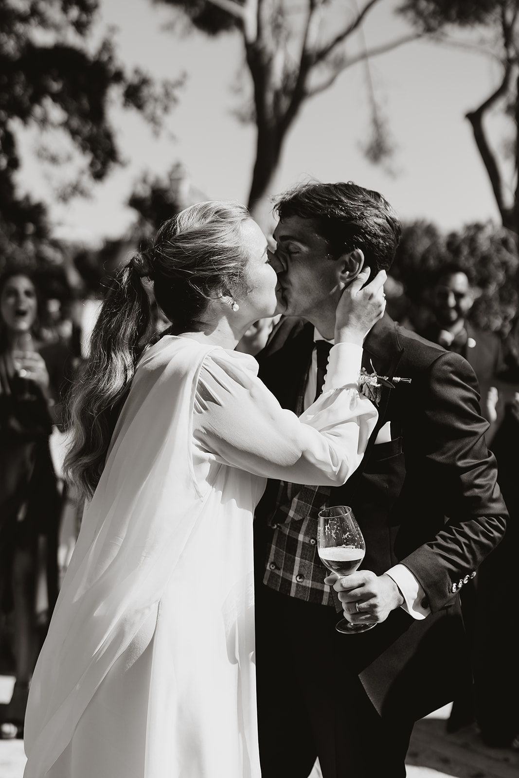 Newlyweds share passionate kiss at outdoor wedding while groom holds champagne glass.