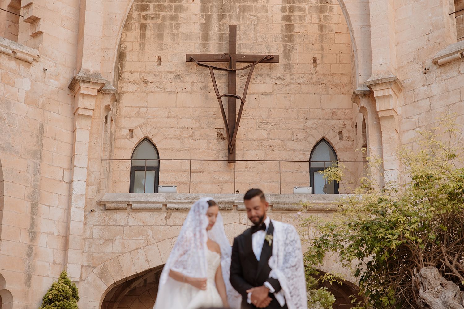 Wedding couple poses in front of stone church wall with gothic windows and crucifix.