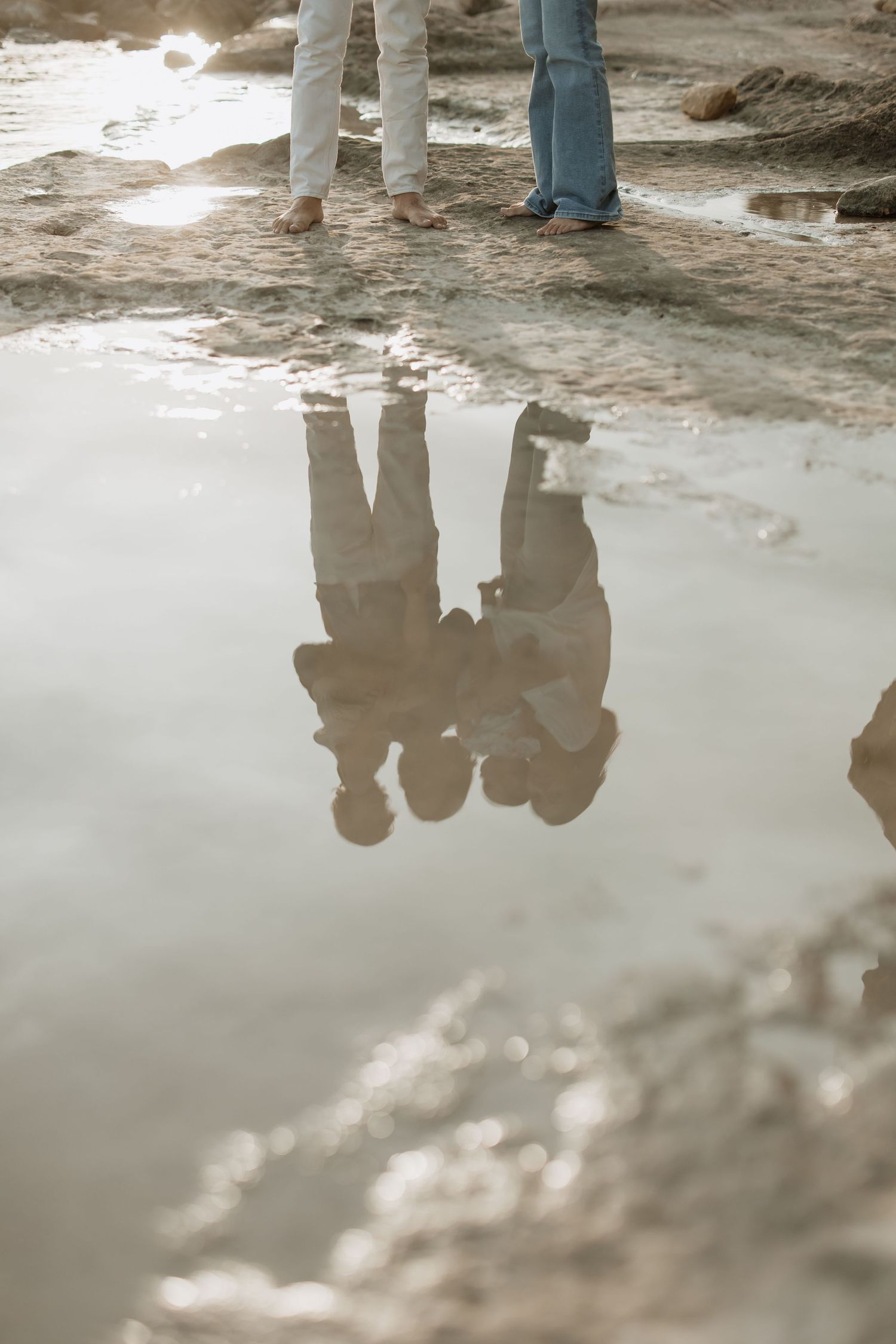 Reflection of figures in a puddle along rocky coastline at sunset.